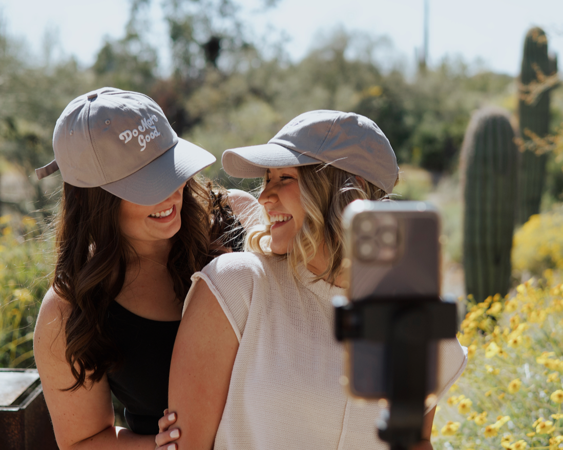 Do More Good Hat, two smiling women wearing gray embroidered caps in a desert setting with cactus and yellow flowers