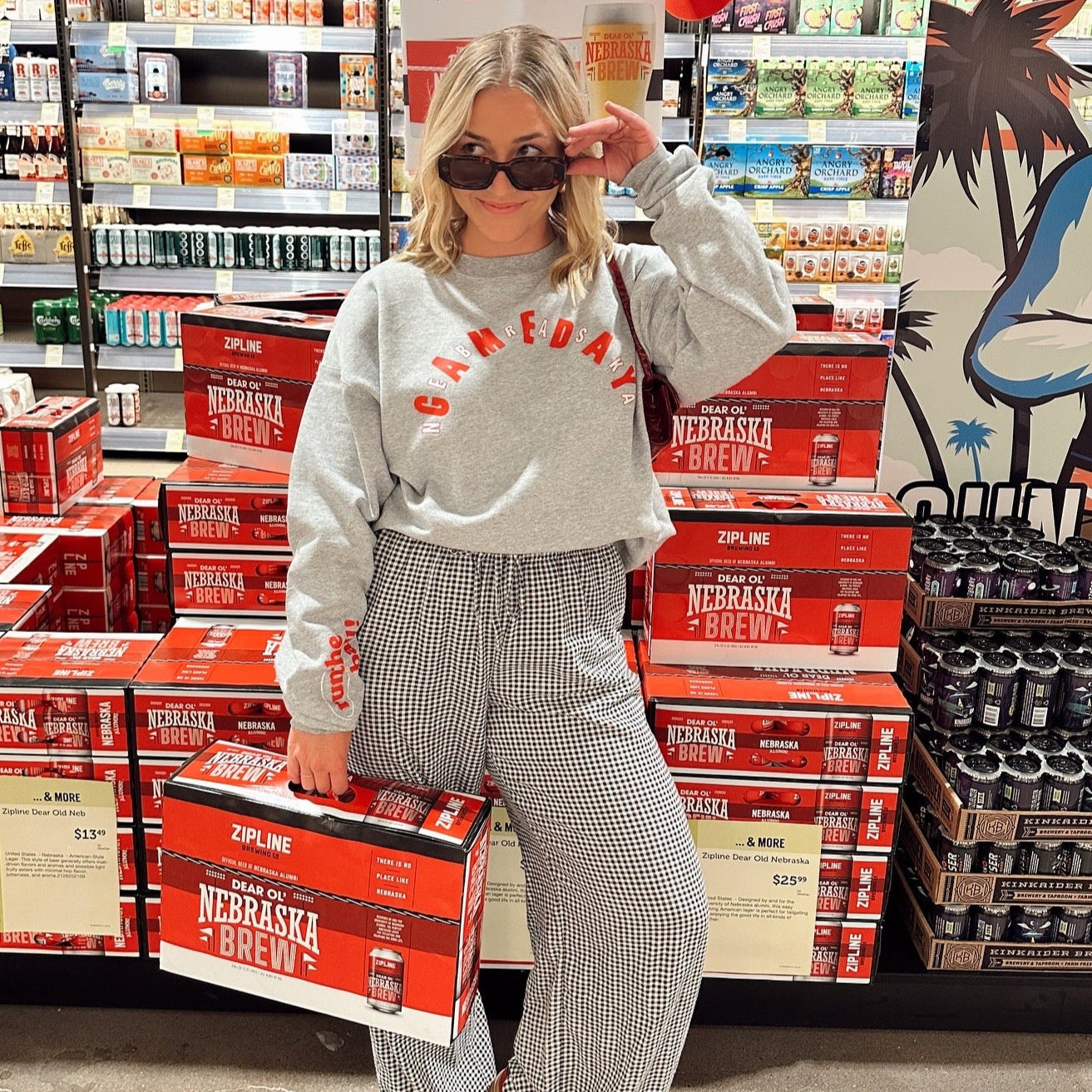 Nebraska Gameday Crew, gray sweatshirt with red arched Gameday text worn by a model in sunglasses in a beer aisle
