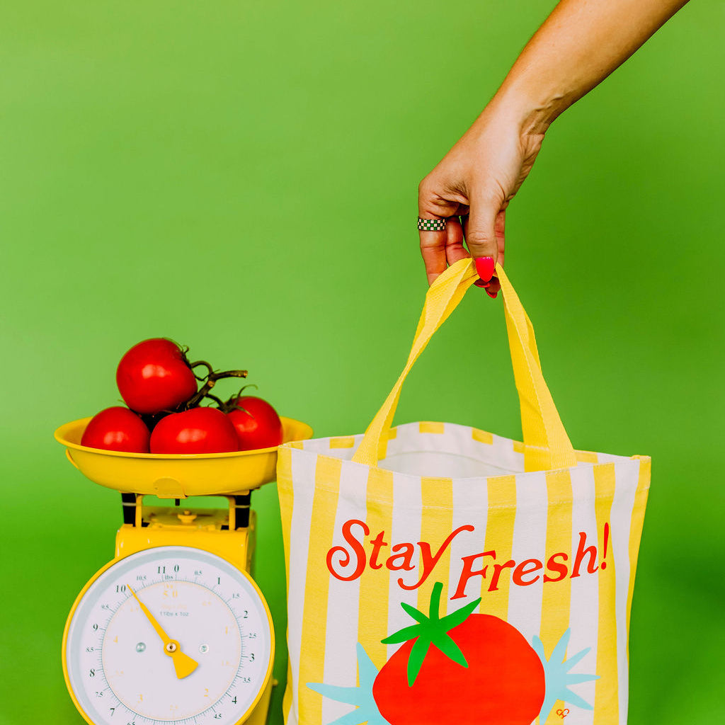 Stay Fresh Tomato Market Tote, yellow striped tote with a red tomato and Stay Fresh text held by a hand beside a scale