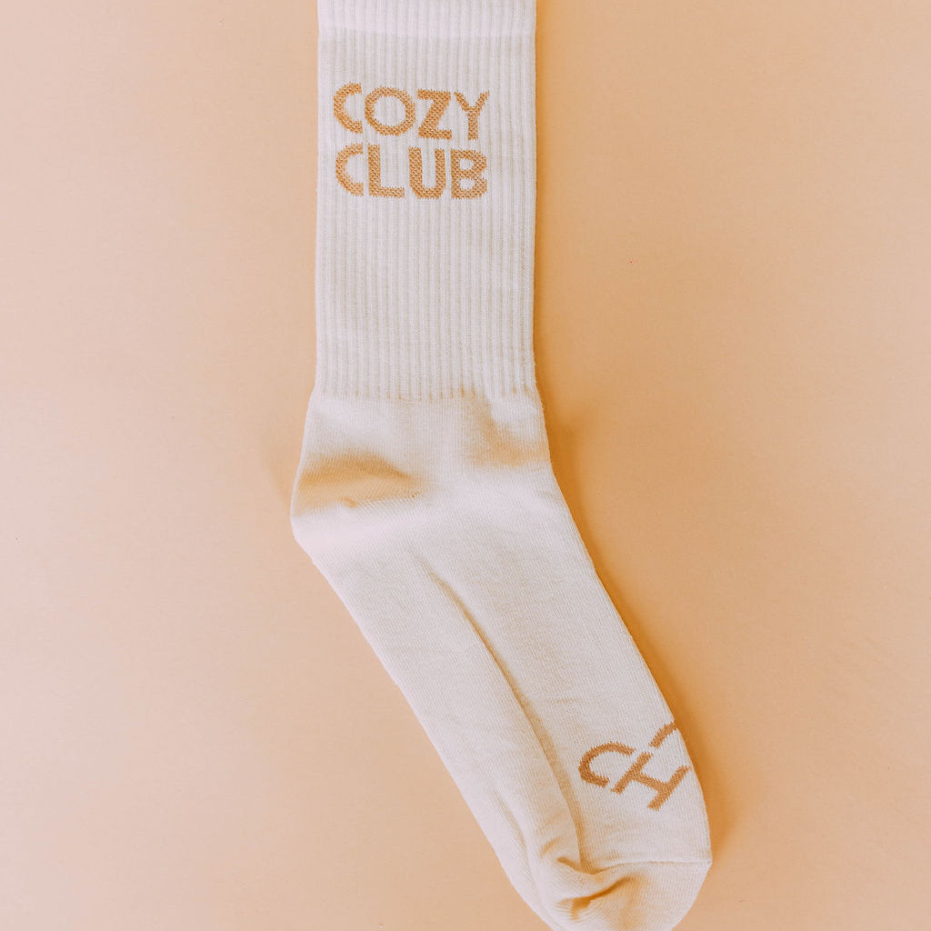 Cozy Club Socks, single cream ribbed crew sock with tan Cozy Club lettering and toe logo on a peach backdrop