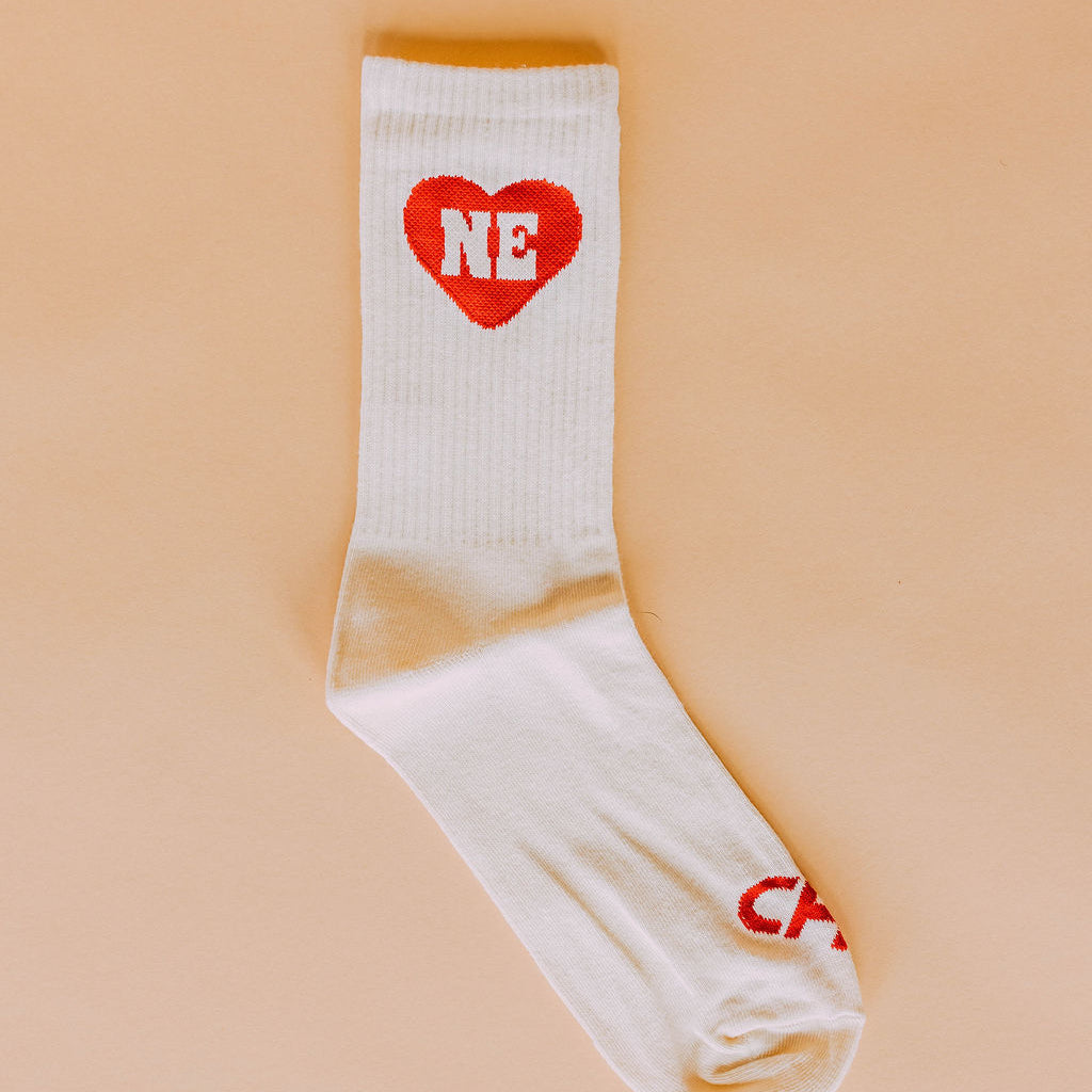 Nebraska Heart Crew Socks, white ribbed crew sock with a red NE heart, flat lay on a peach background