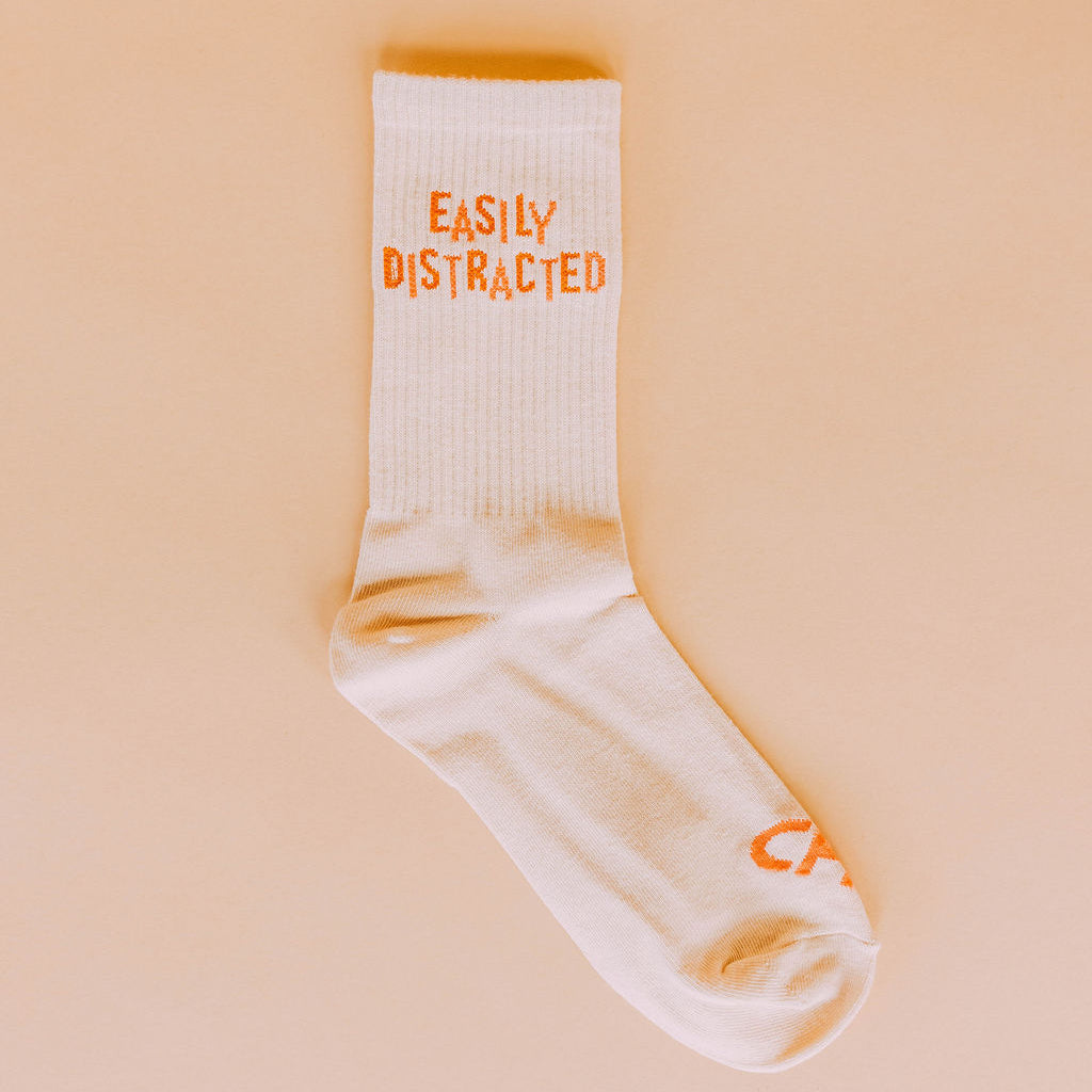 Easily Distracted Socks, single cream crew sock with orange Easily Distracted text, flat lay on peach background