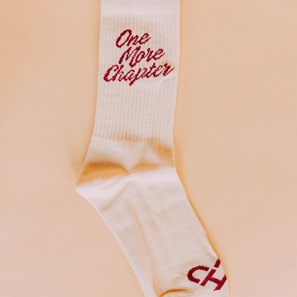 One More Chapter Socks, single cream crew sock with red One More Chapter script, flat lay on peach background