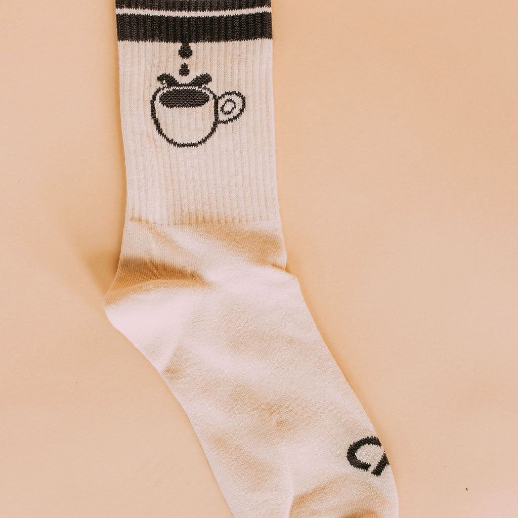 Coffee Cup Drip Socks, cream crew sock with black stripes and a dripping coffee cup graphic, flat lay on peach