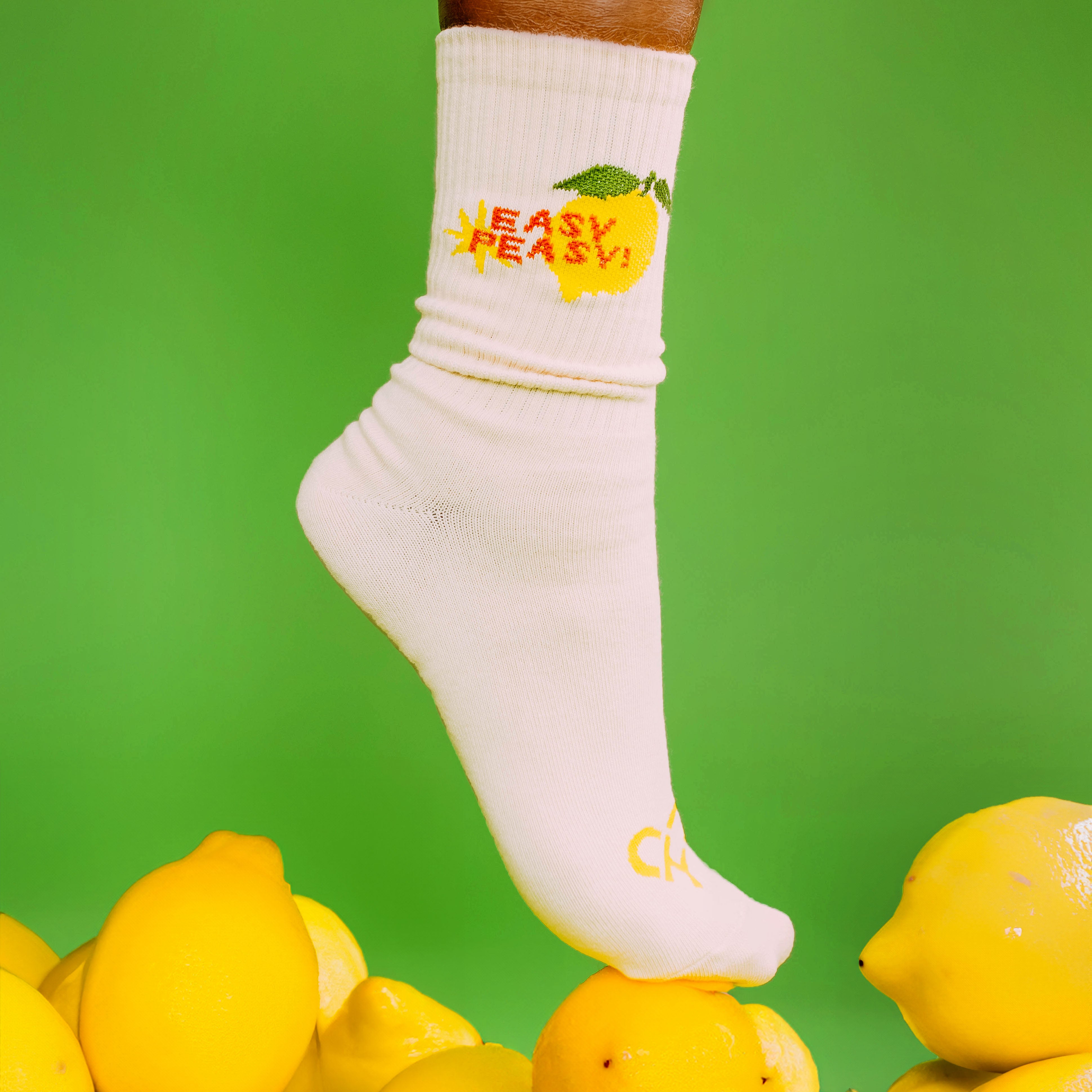 Easy Peasy Lemon Crew Socks, cream sock with a lemon and red Easy Peasy text worn on a foot atop a pile of lemons