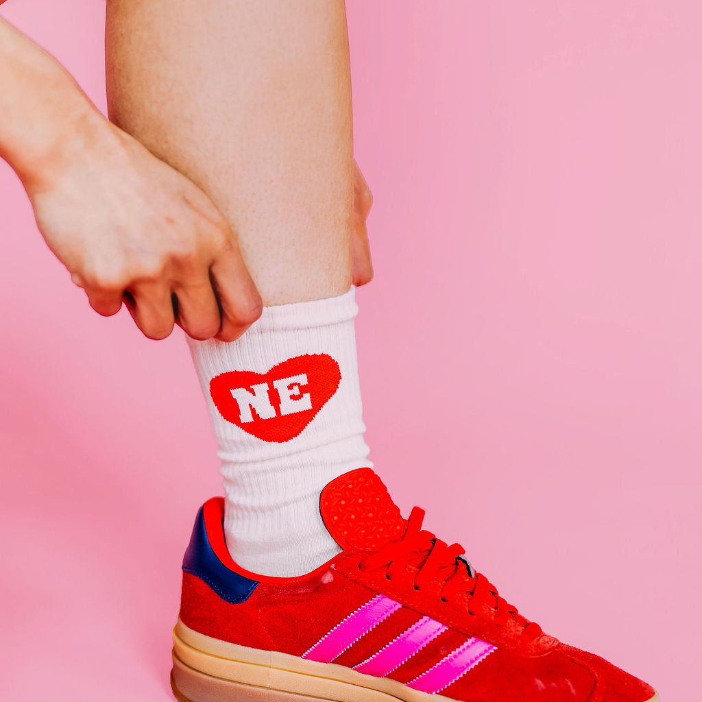 Nebraska Heart Crew Socks, white crew sock with red heart and white NE worn with red sneakers on a pink backdrop