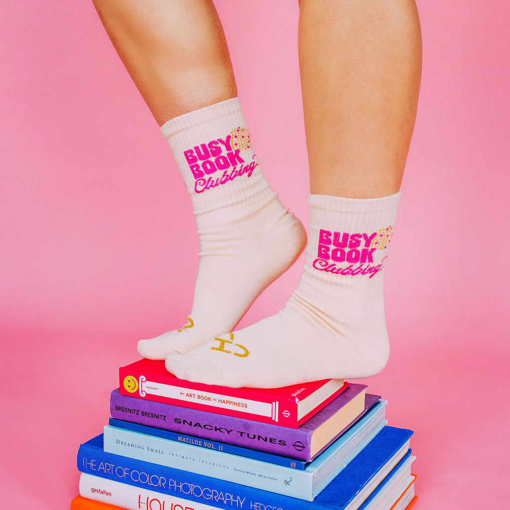 Busy Book Clubbing Socks, cream socks with pink Busy Book Clubbing text worn by a model standing on a stack of books
