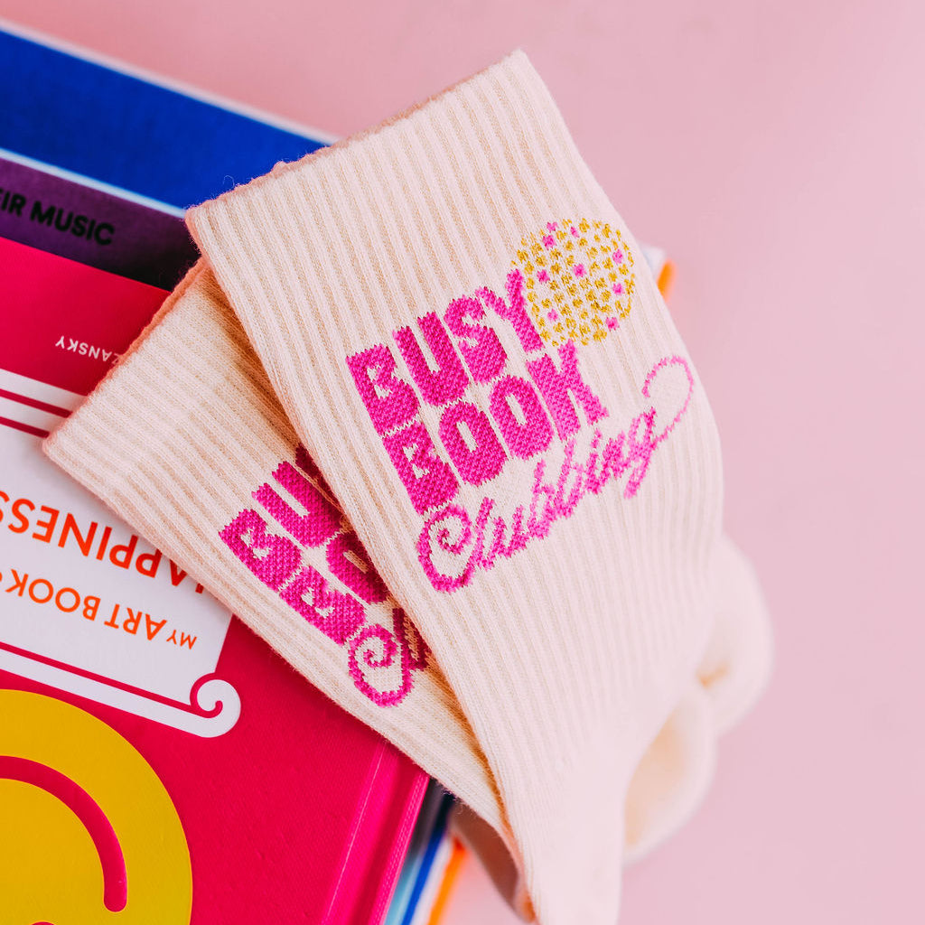 Busy Book Clubbing Socks, cream socks with pink Busy Book Clubbing text and a disco ball, draped on a stack of books