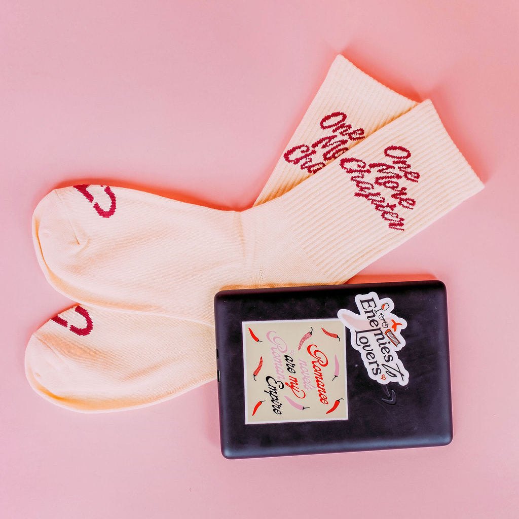One More Chapter Socks, cream socks with red One More Chapter script beside an e reader with stickers, on pink