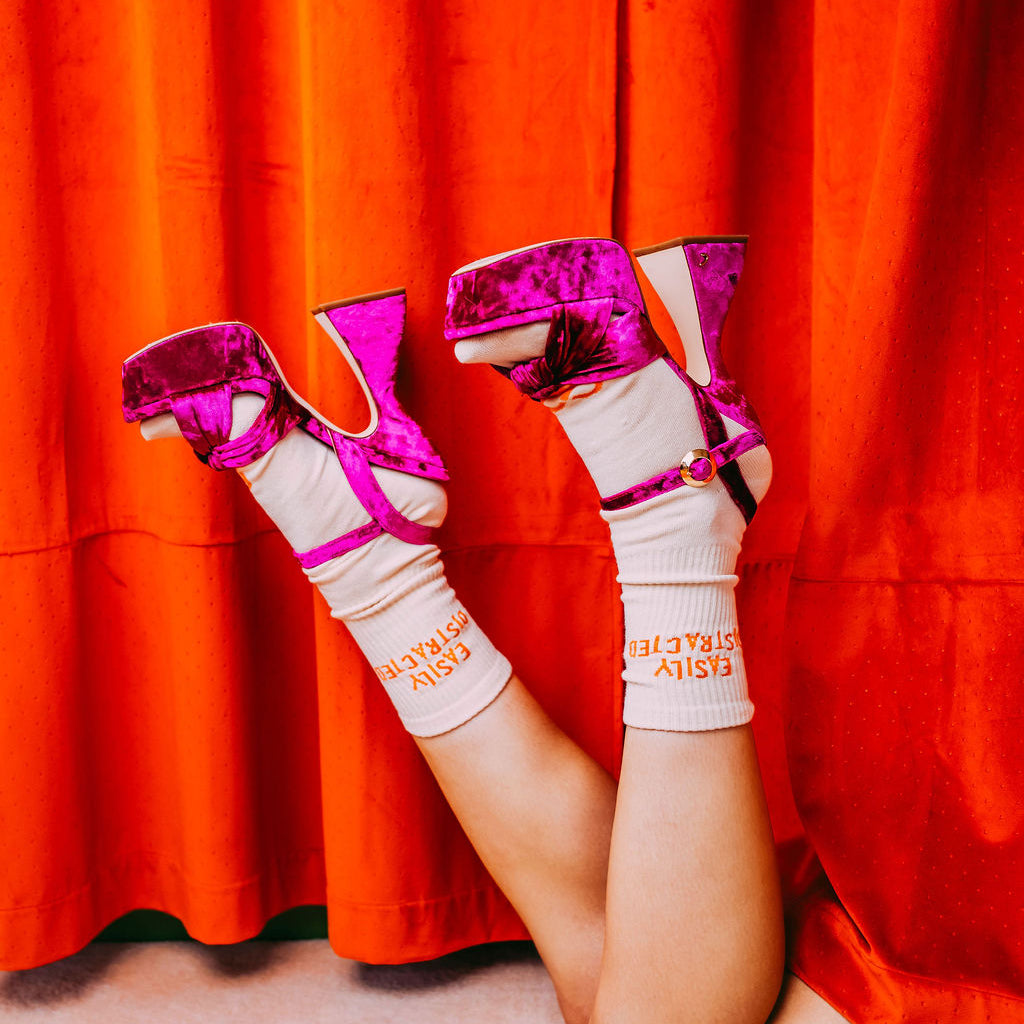 Easily Distracted Socks, cream socks with orange text worn with pink metallic heels, legs raised by a red curtain