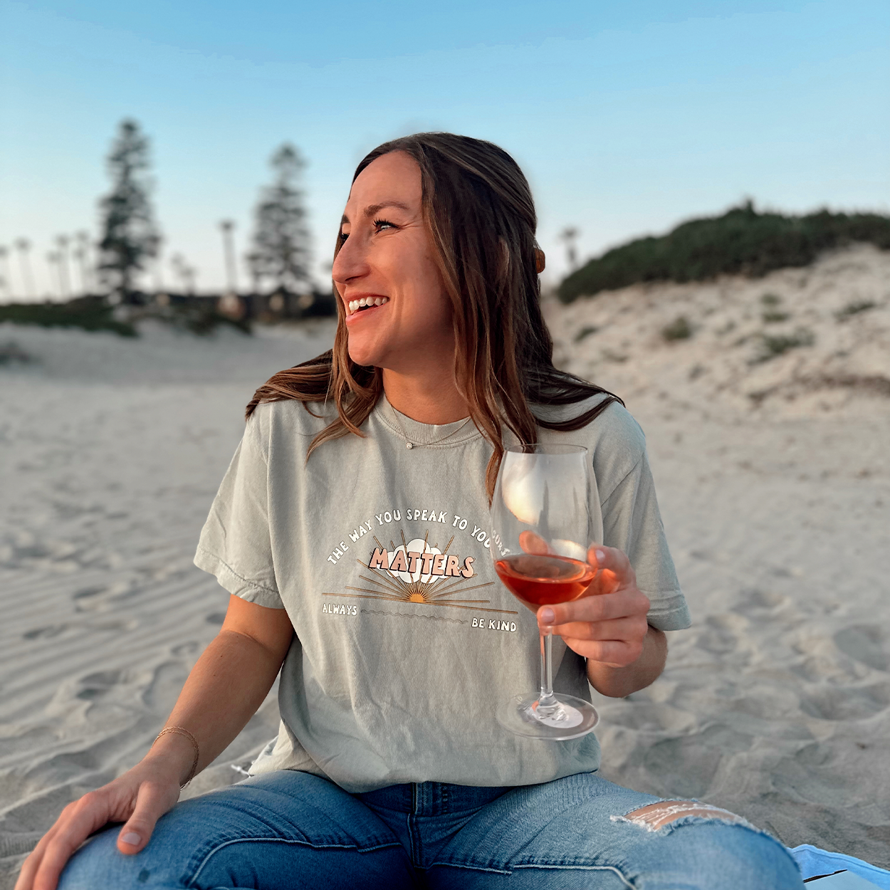 The Way You Speak To Yourself Matters Tee in sage, worn by a smiling model sitting on a beach holding a glass of rose