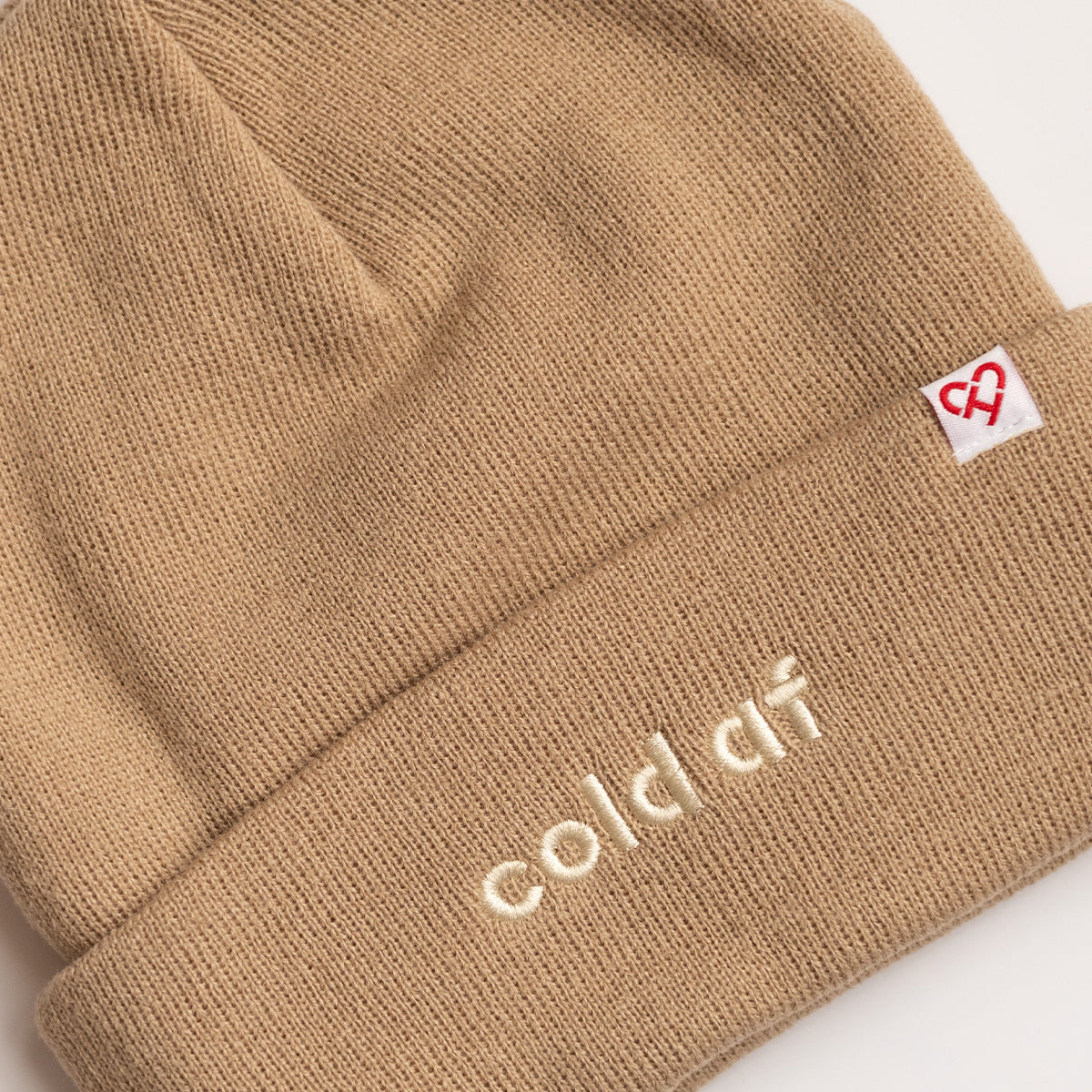 Cold AF Beanie, close up of tan knit cuff with cream embroidered cold af text and a red heart logo tag
