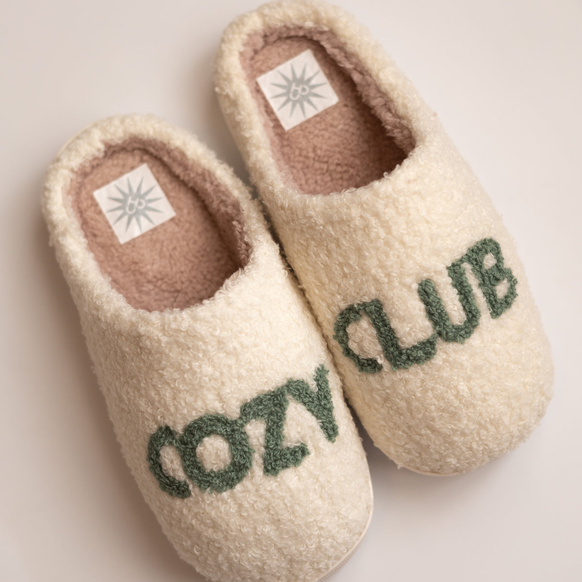 Cozy Club Slippers, cream sherpa slippers with green tufted Cozy Club lettering split across the pair, top view
