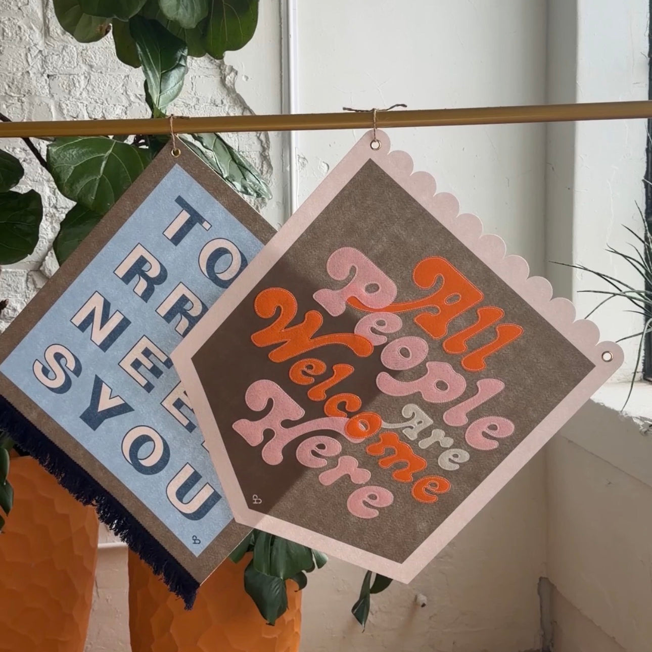 All People Are Welcome Here Pennant, olive felt banner with pink and orange script hung on a brass rod by plants