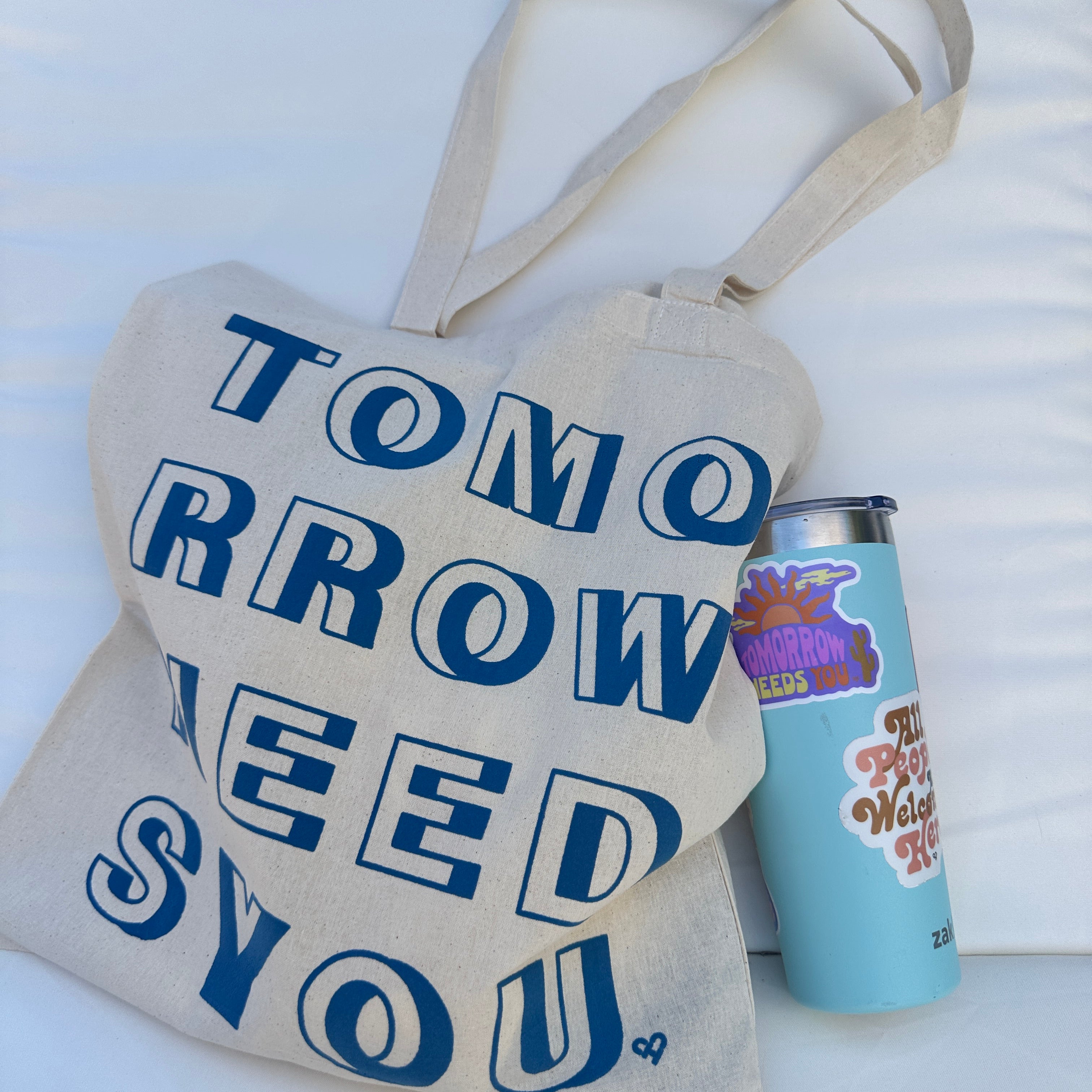 Tomorrow Needs You Sticker on a light blue tumbler beside a canvas tote printed with blue Tomorrow Needs You text