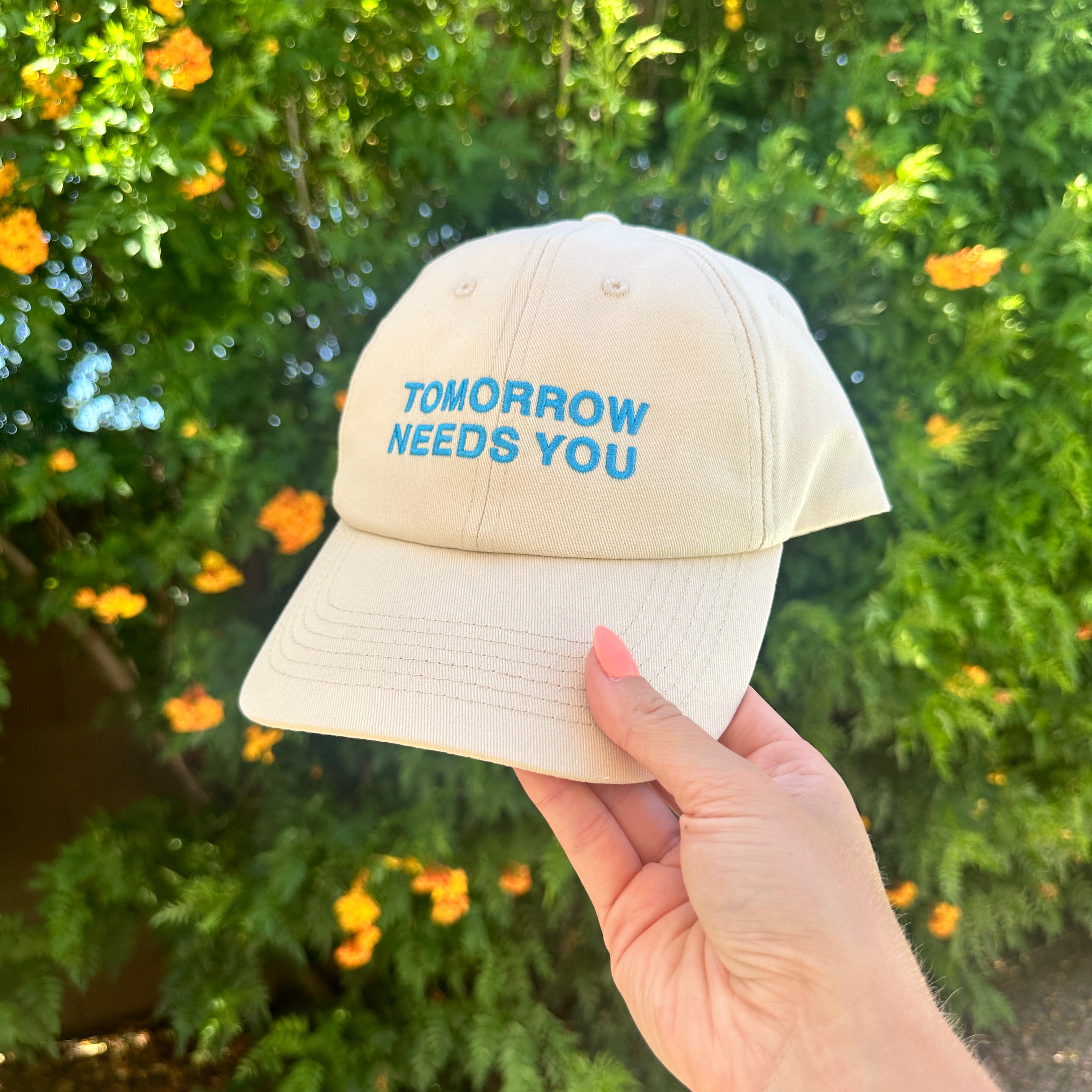 Tomorrow Needs You Hat, cream dad cap with blue embroidered Tomorrow Needs You text held up by orange flowers