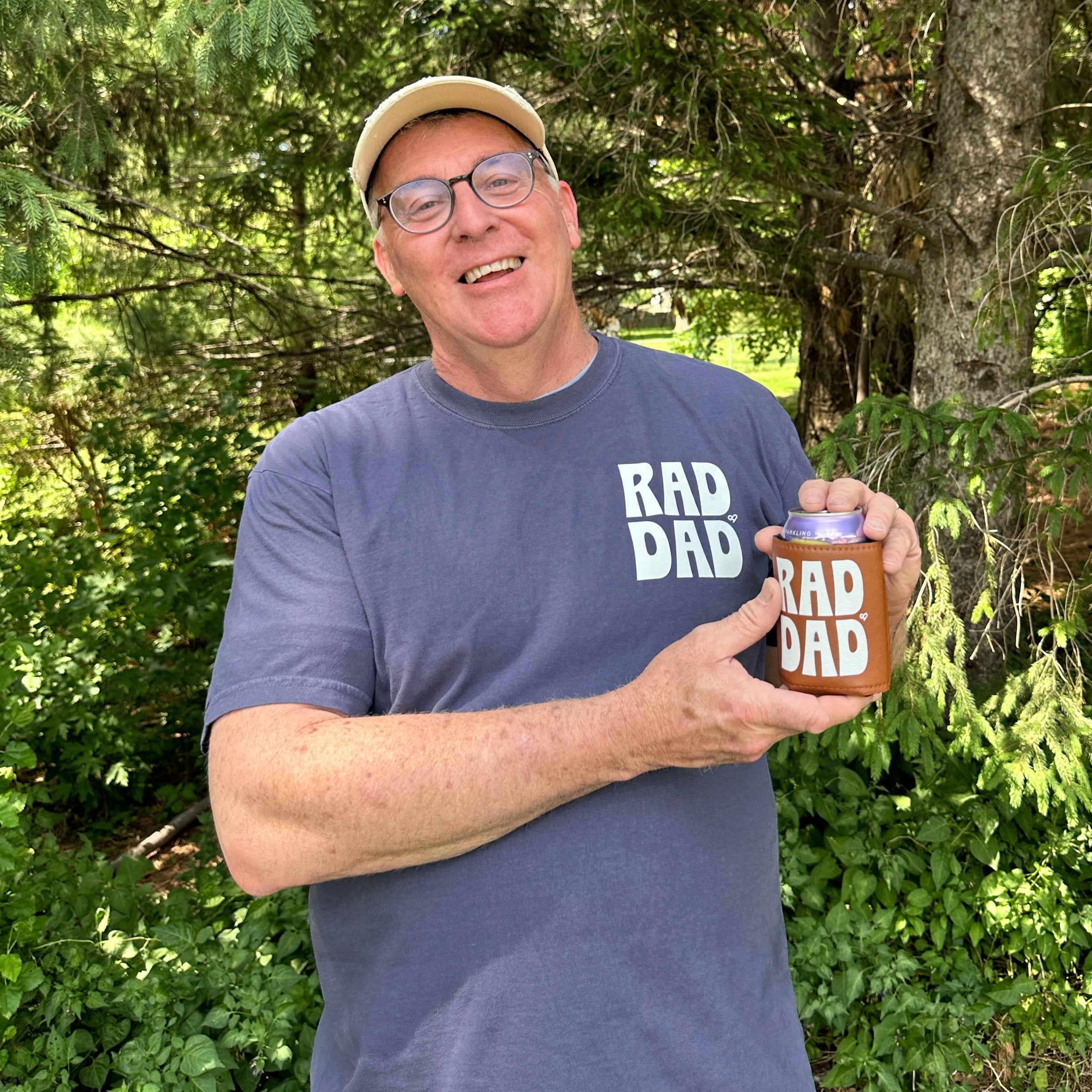Rad Dad Tee worn by a smiling man outdoors among trees, holding a matching brown Rad Dad koozie