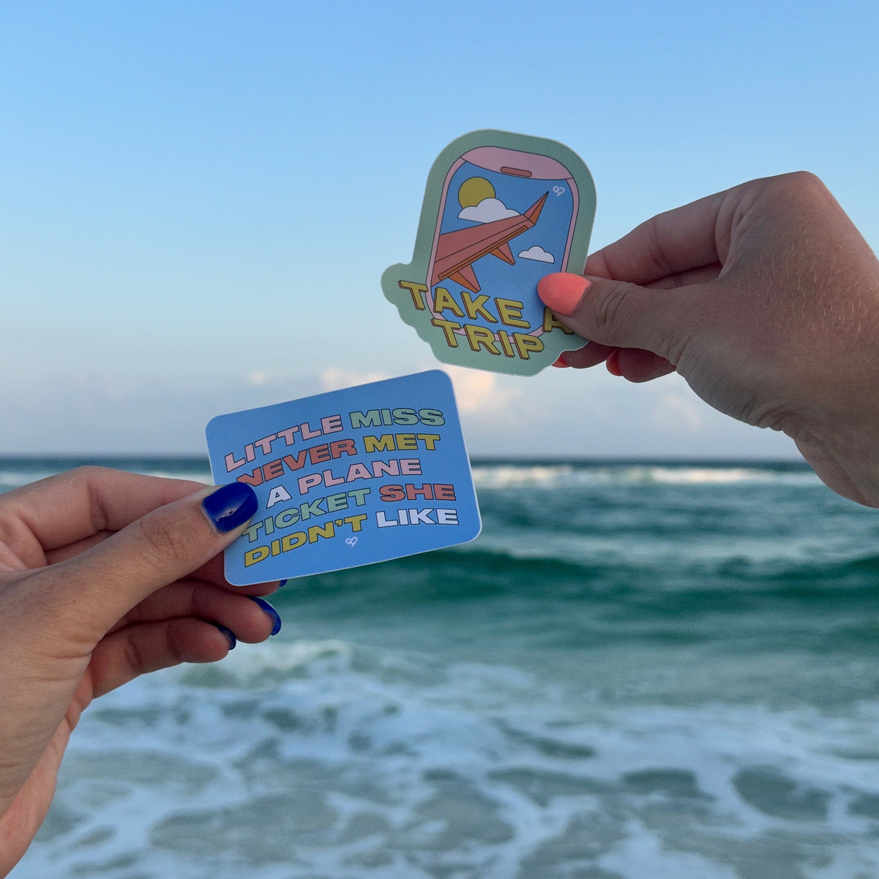 Plane Ticket Sticker and Take A Trip Sticker held up by two hands in front of ocean waves at the beach