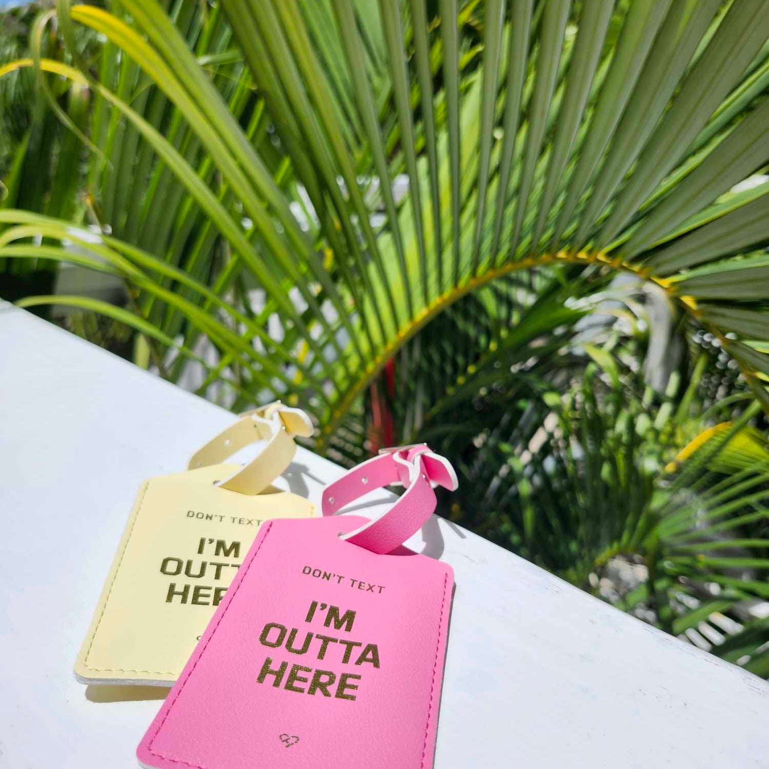 Don't Text Luggage Tag in yellow and pink with gold Don't Text I'm Outta Here text on a white ledge by palm fronds