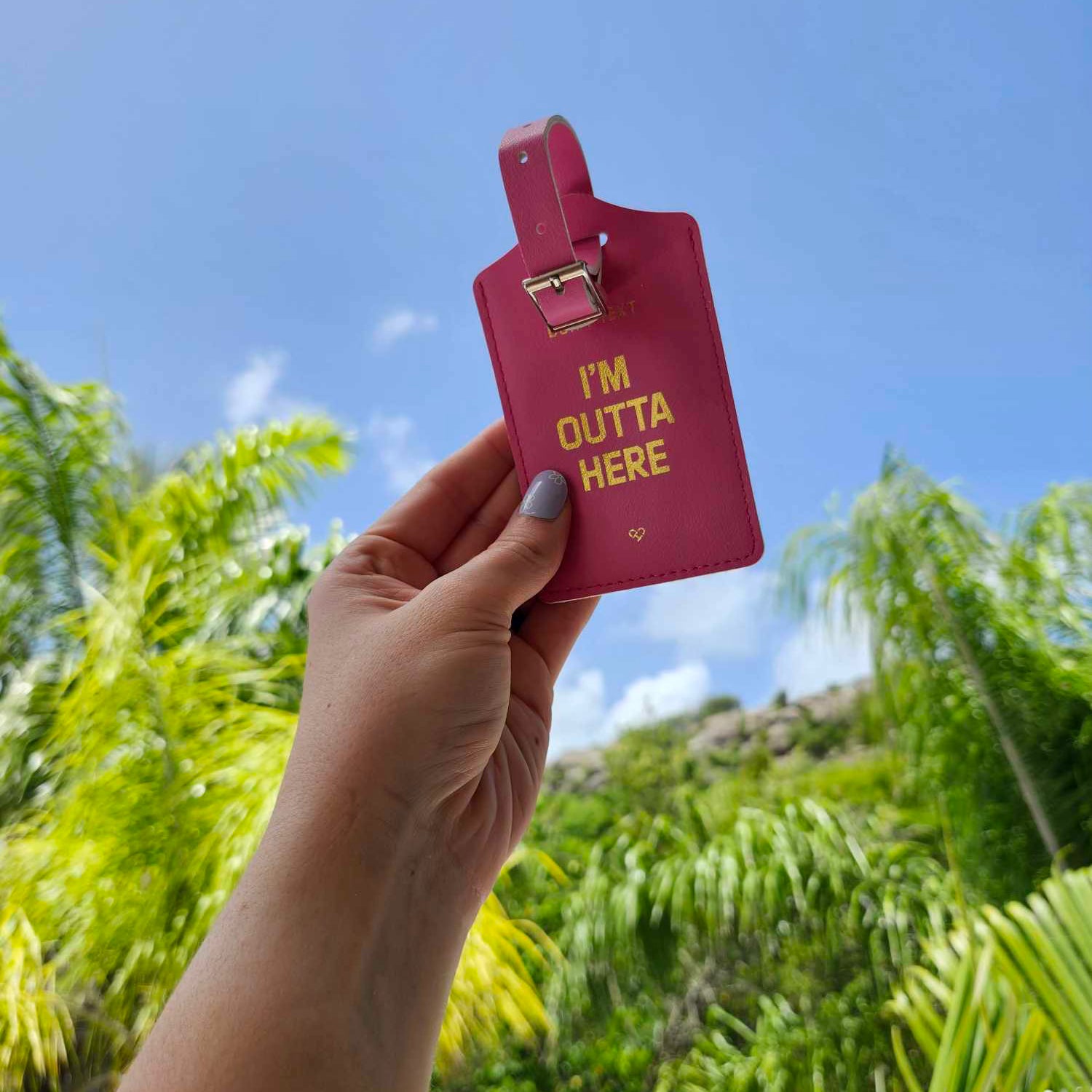 Don't Text Luggage Tag in pink with gold I'm Outta Here text and buckle strap held up against blue sky and palms