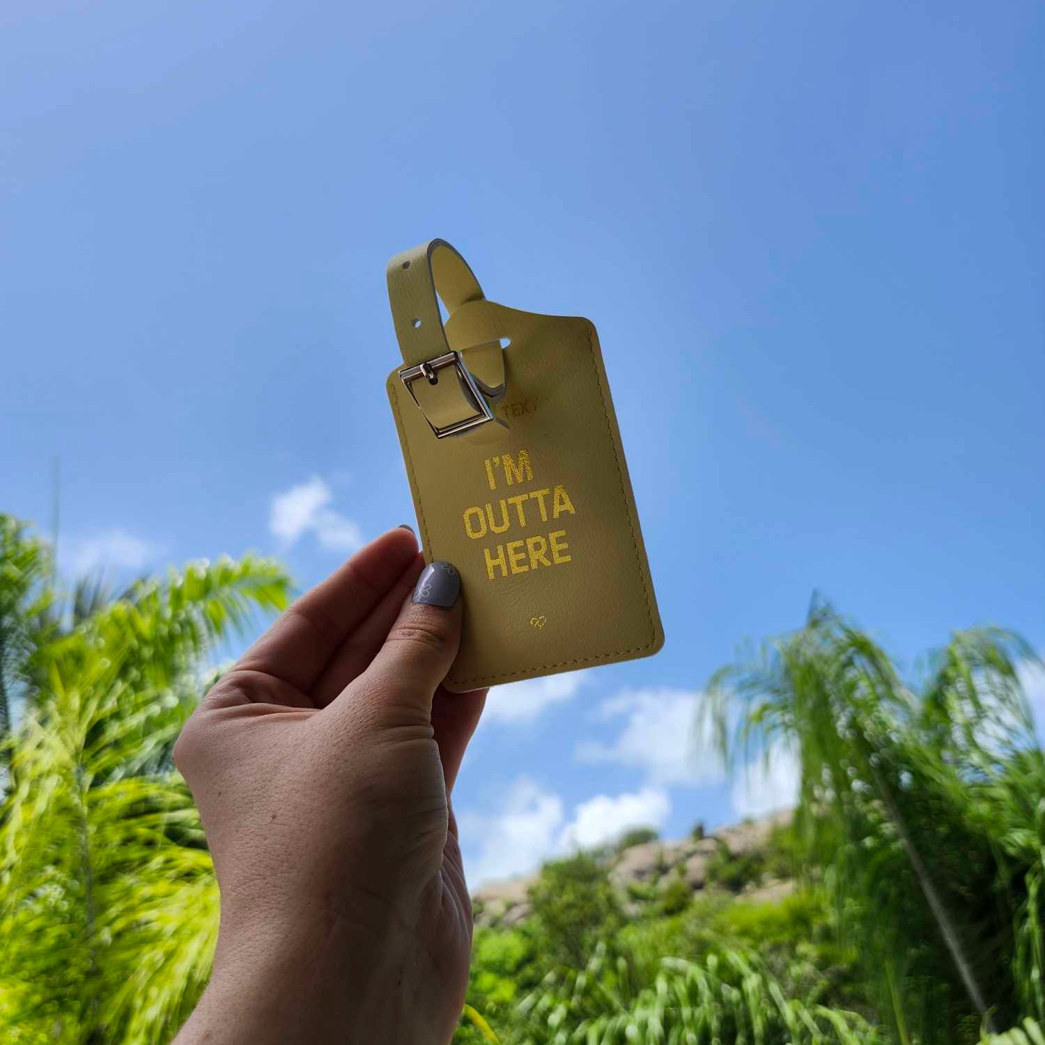 Don't Text Luggage Tag in yellow with gold I'm Outta Here text and buckle strap held up against sky and palms