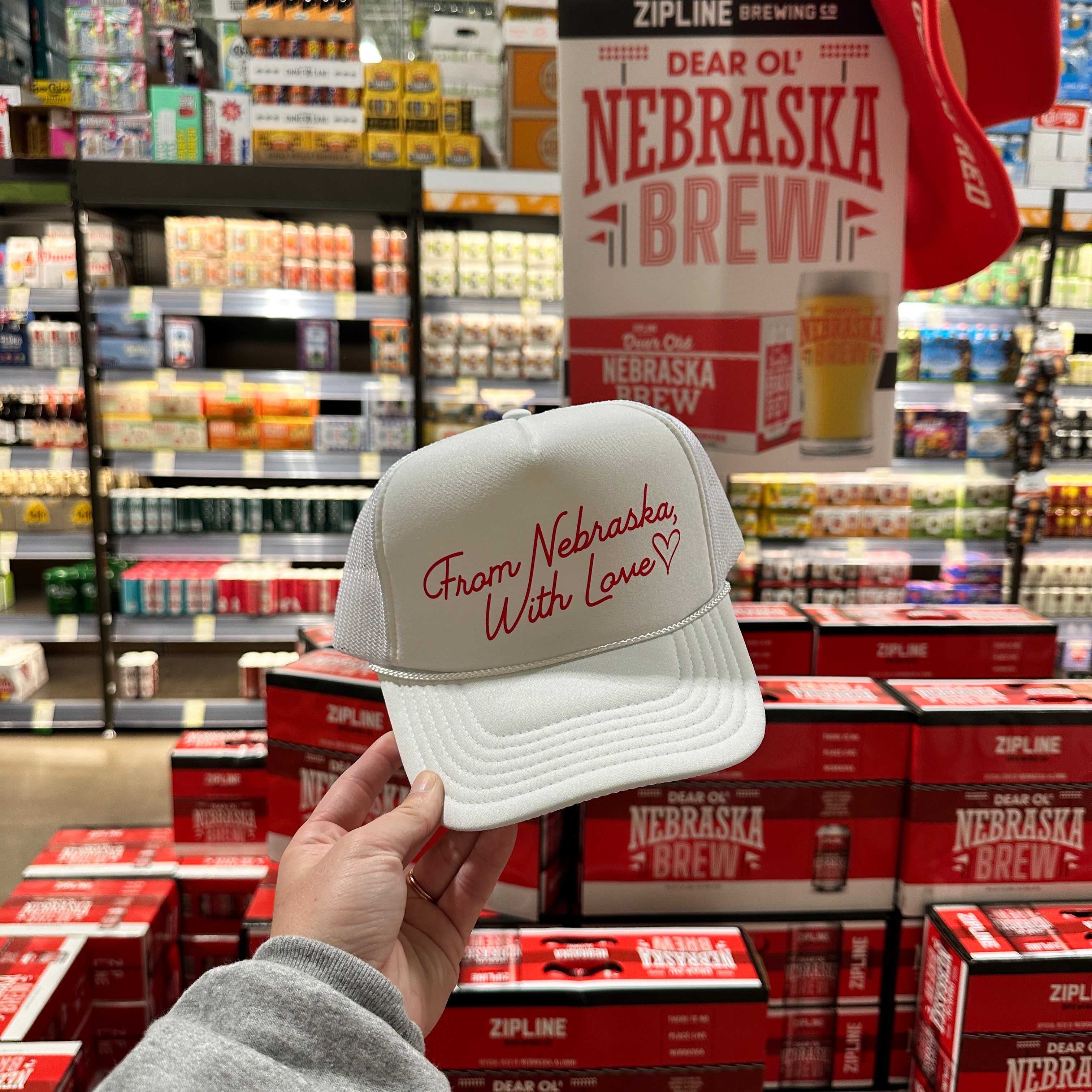 From Nebraska With Love Trucker Hat held in a store aisle in front of stacked Nebraska Brew beer cases