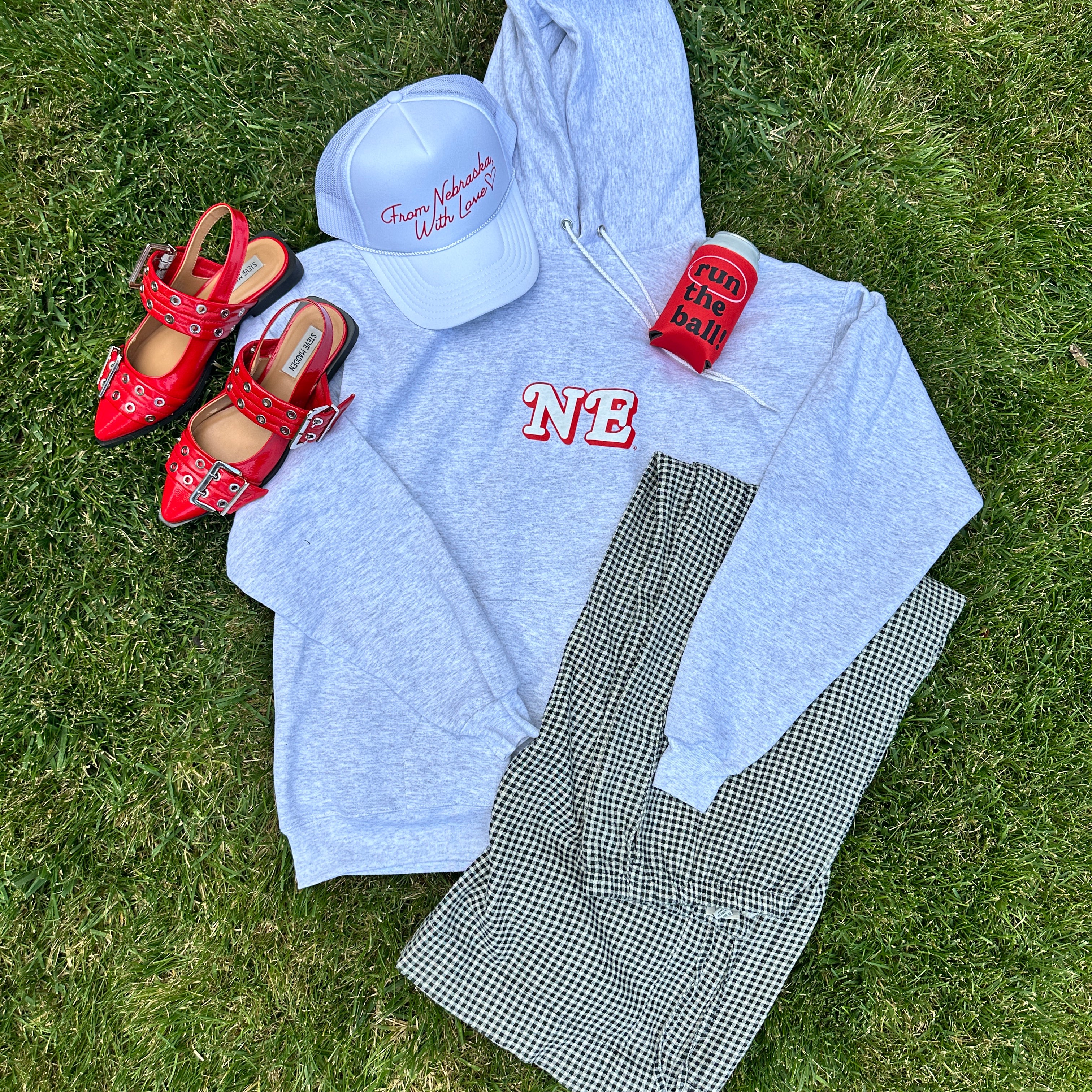 Run The Ball Koozie in a flat lay on grass with a gray NE hoodie, white trucker hat, red flats and gingham pants