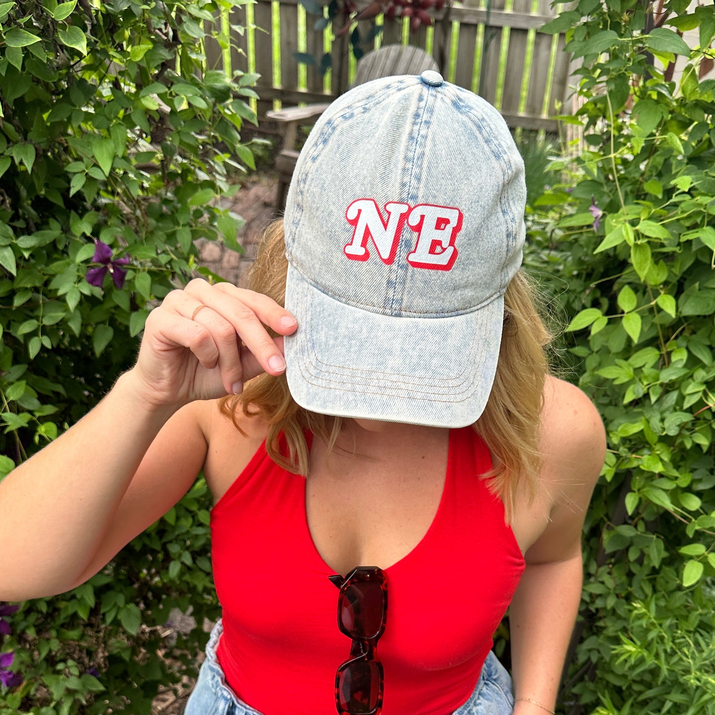 NE Denim Hat worn by a model in a red tank top and jeans, light wash denim cap with red and white NE in a garden