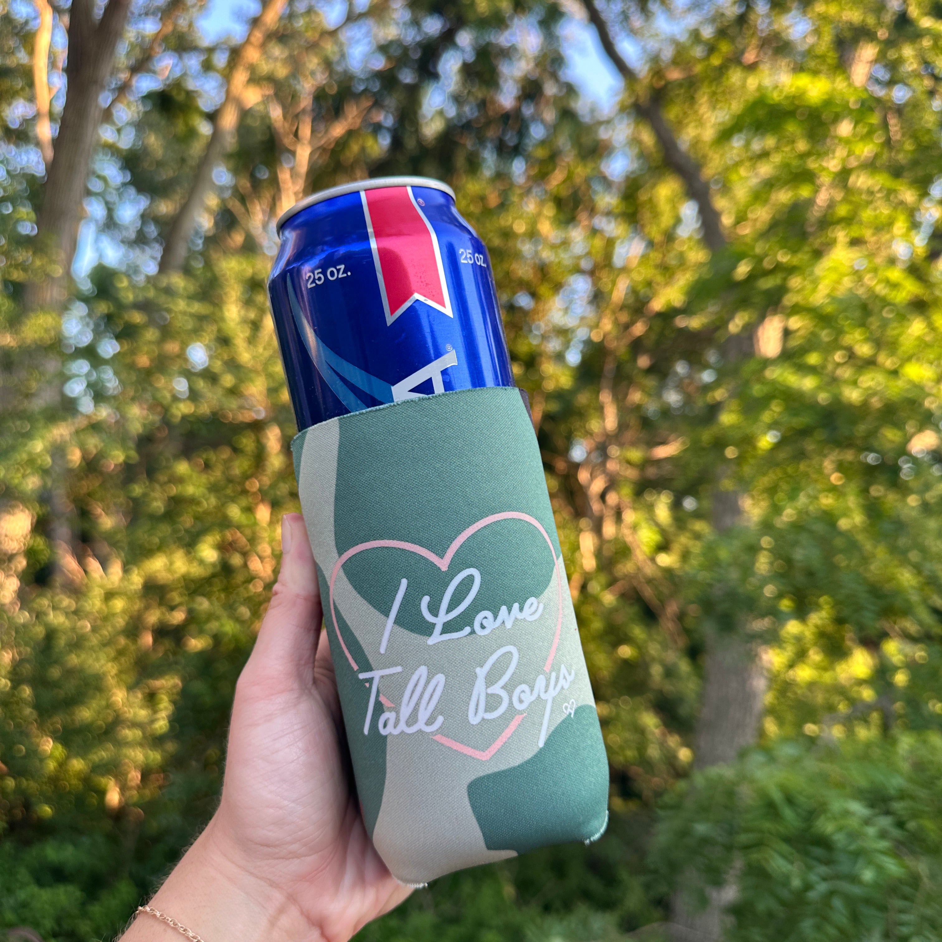 I Love Tall Boys Camo Koozie, green camo sleeve with pink heart and script I Love Tall Boys held up against trees