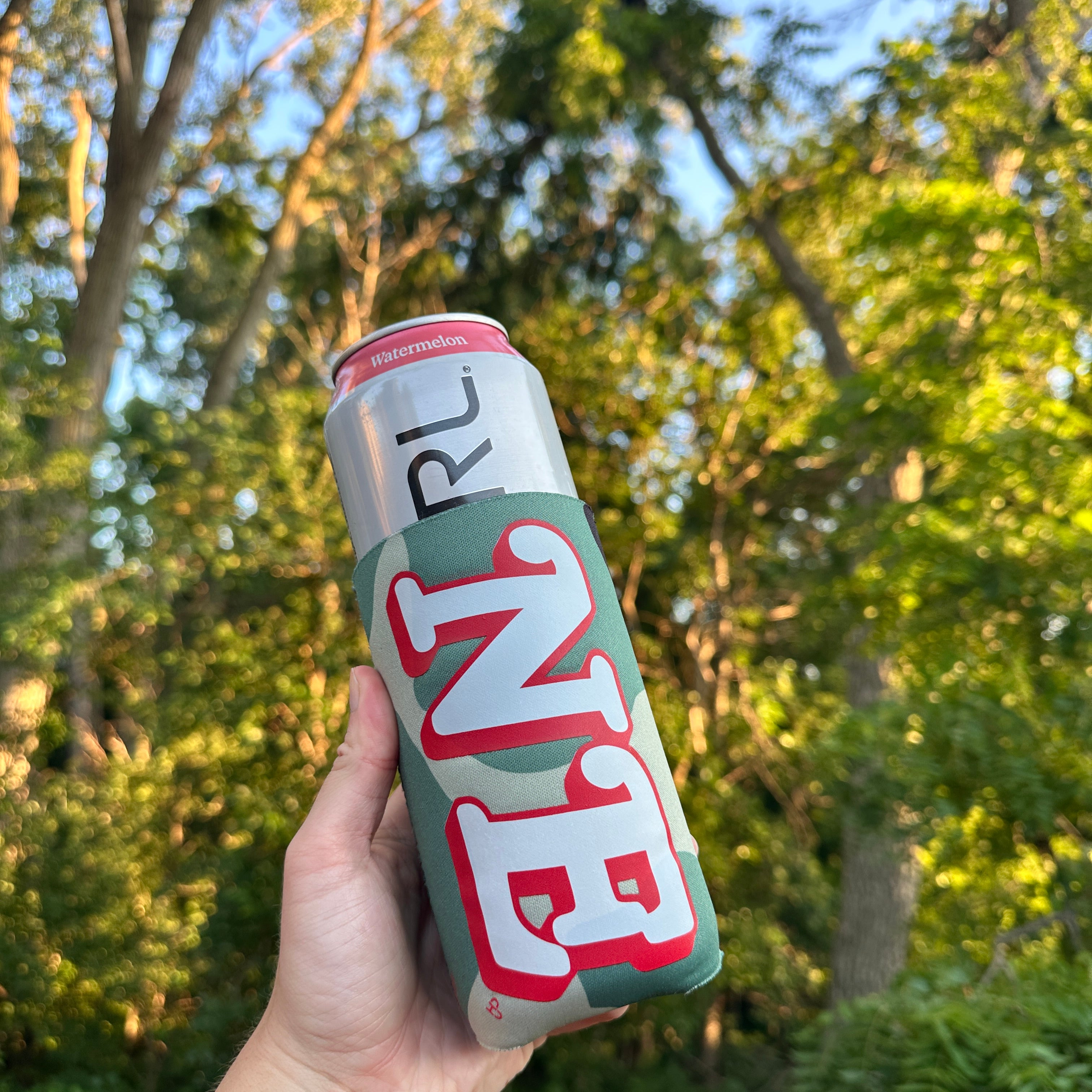 NE Camo Tall Boy Koozie, green camo sleeve with white and red NE letters on a tall can, held up against trees