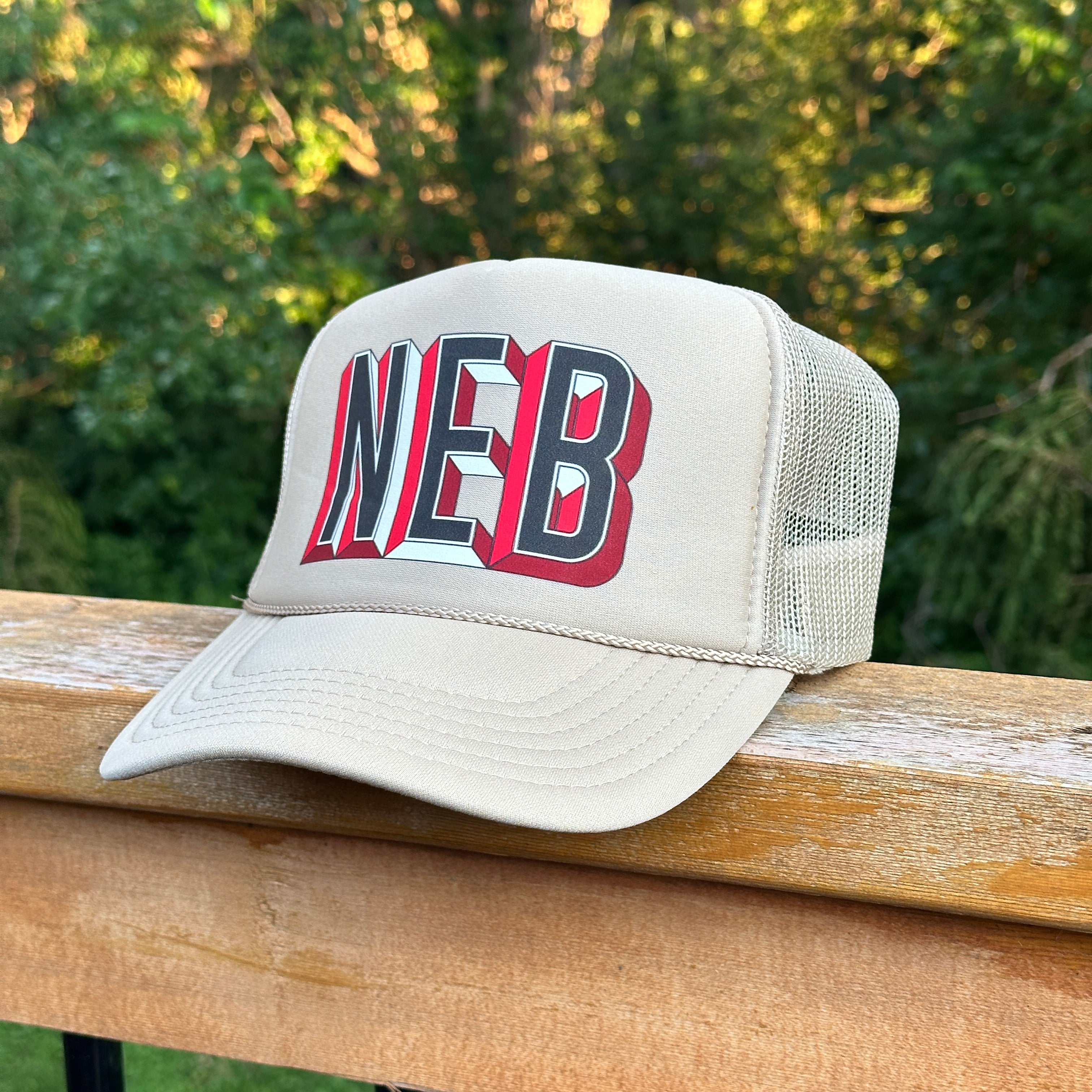 NEB Trucker Hat, tan foam trucker with black, white and red 3D NEB letters and mesh back on a wood railing