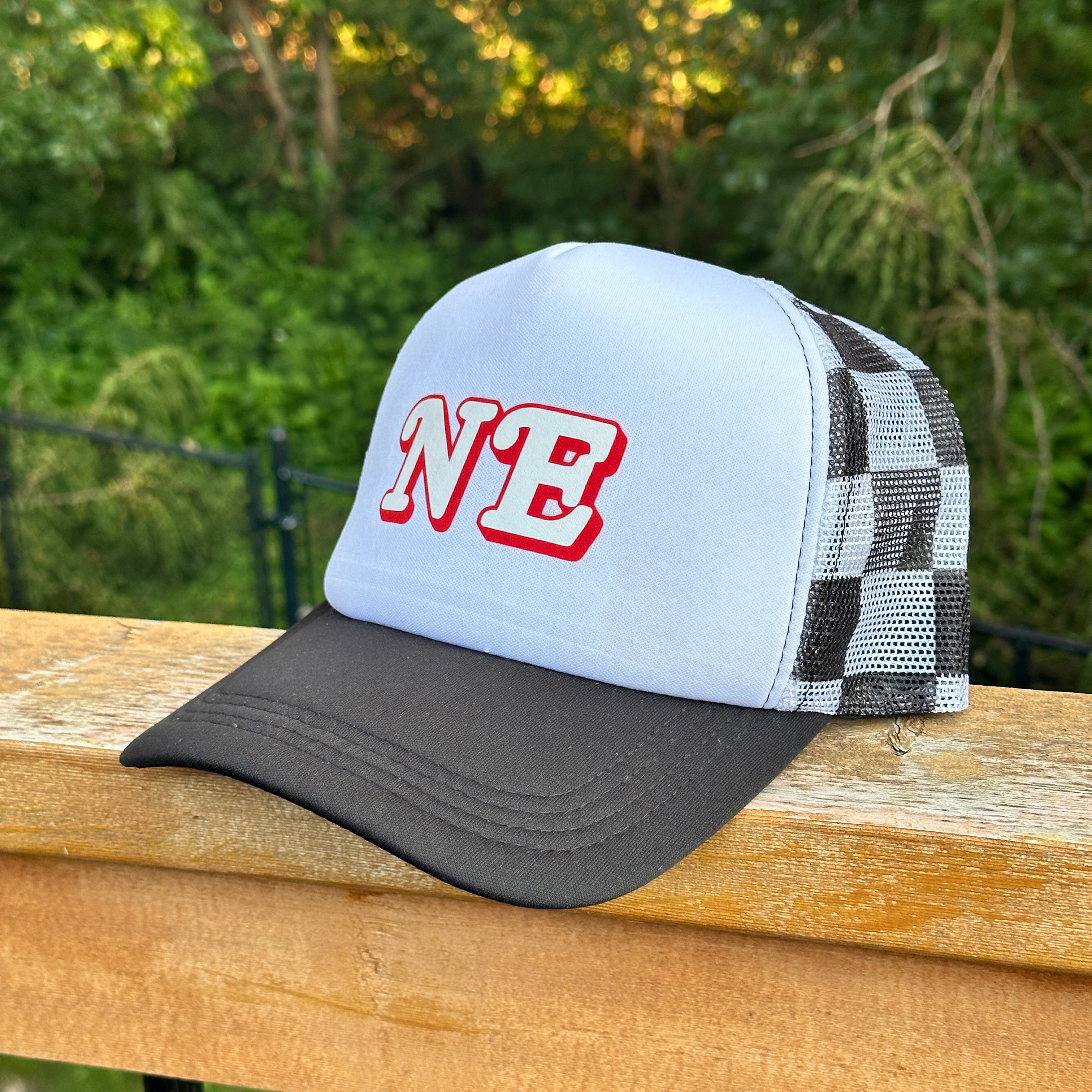 NE Checkered Trucker Hat, white foam front with red NE letters, black brim and checkered mesh back on a wood railing
