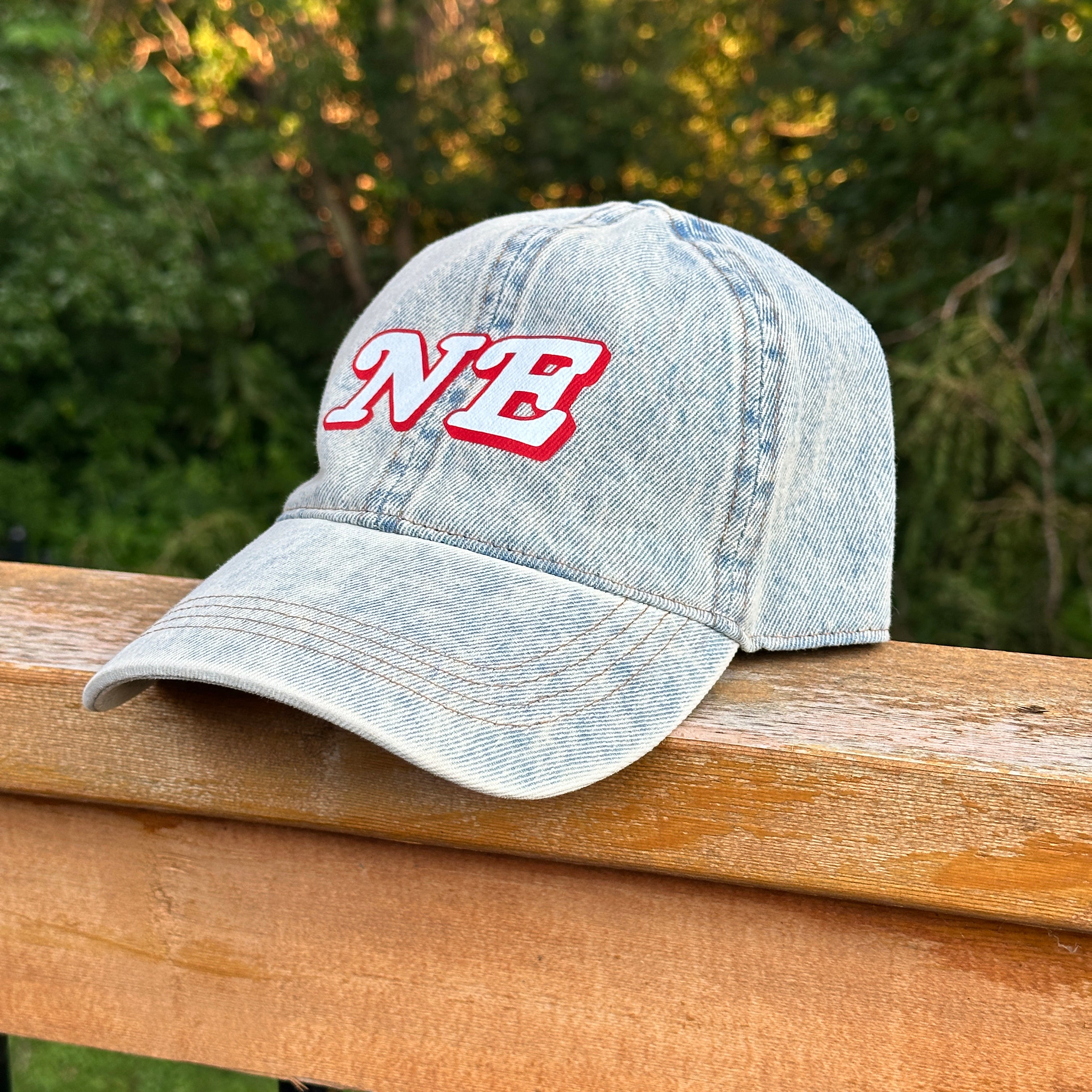 NE Denim Hat, light acid wash denim baseball cap with white and red NE letters resting on a wood deck railing