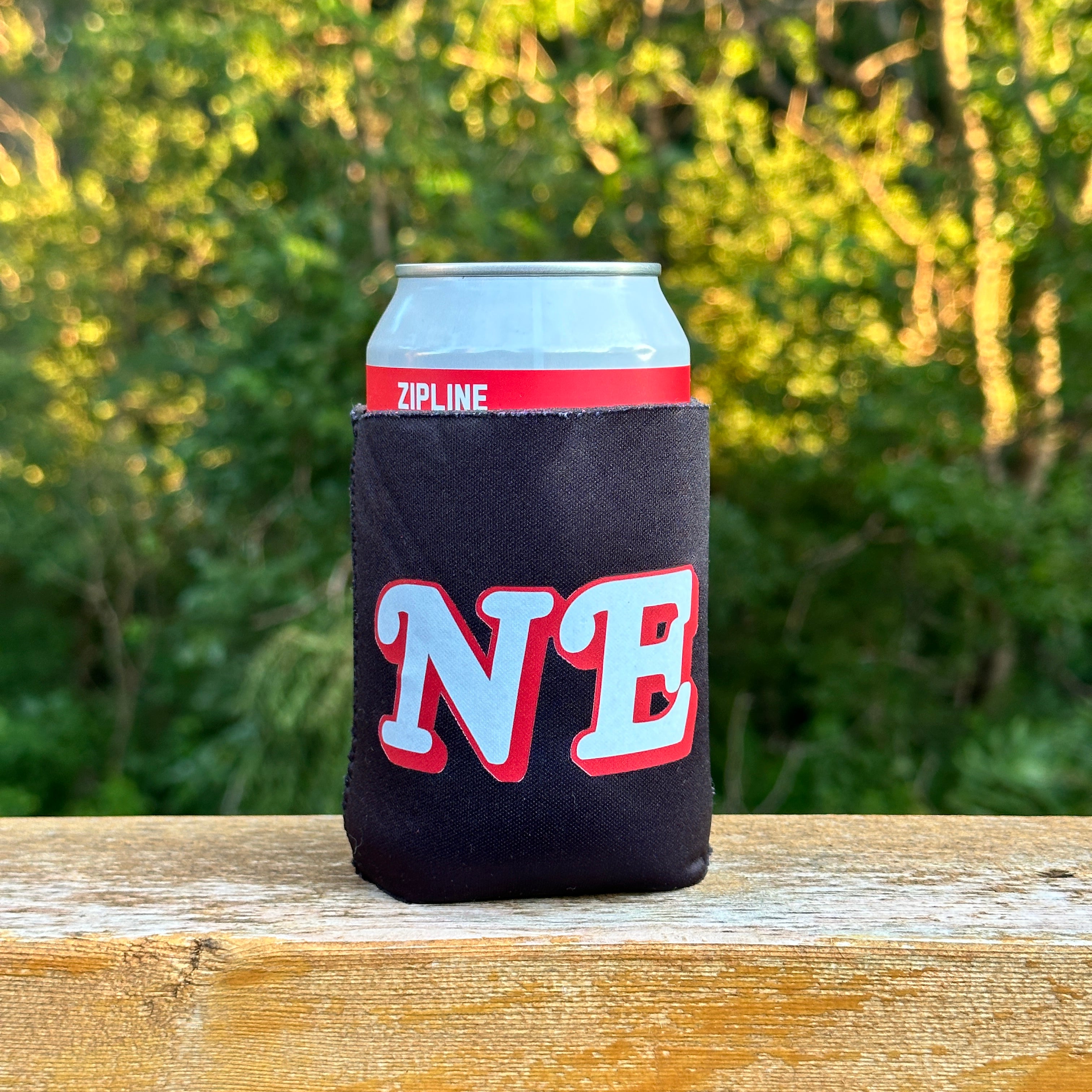 NE Black Koozie, black can koozie with white and red NE letters on a can resting on a wood railing