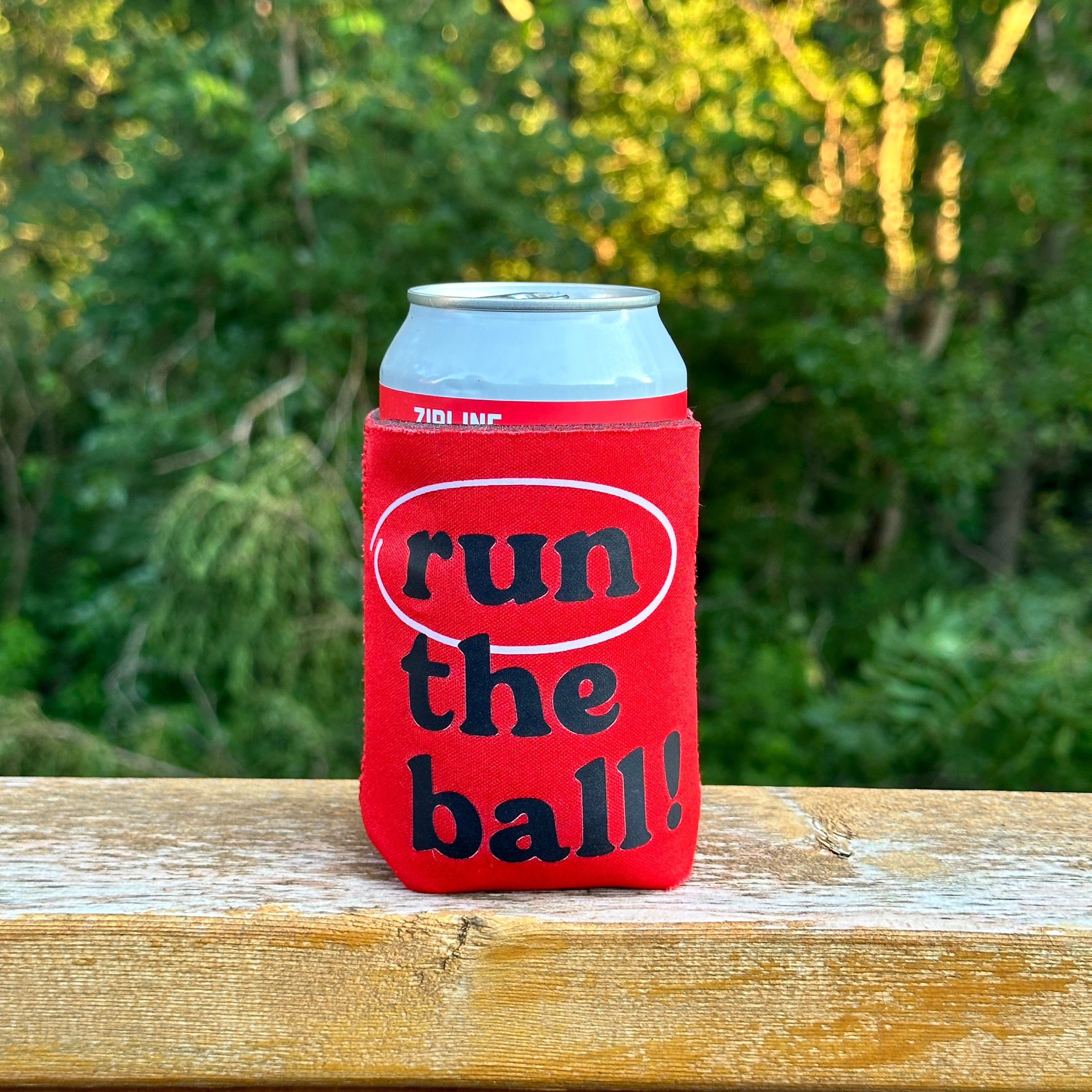 Run The Ball Koozie, red can koozie with black lowercase run the ball! text on a can on a wood railing
