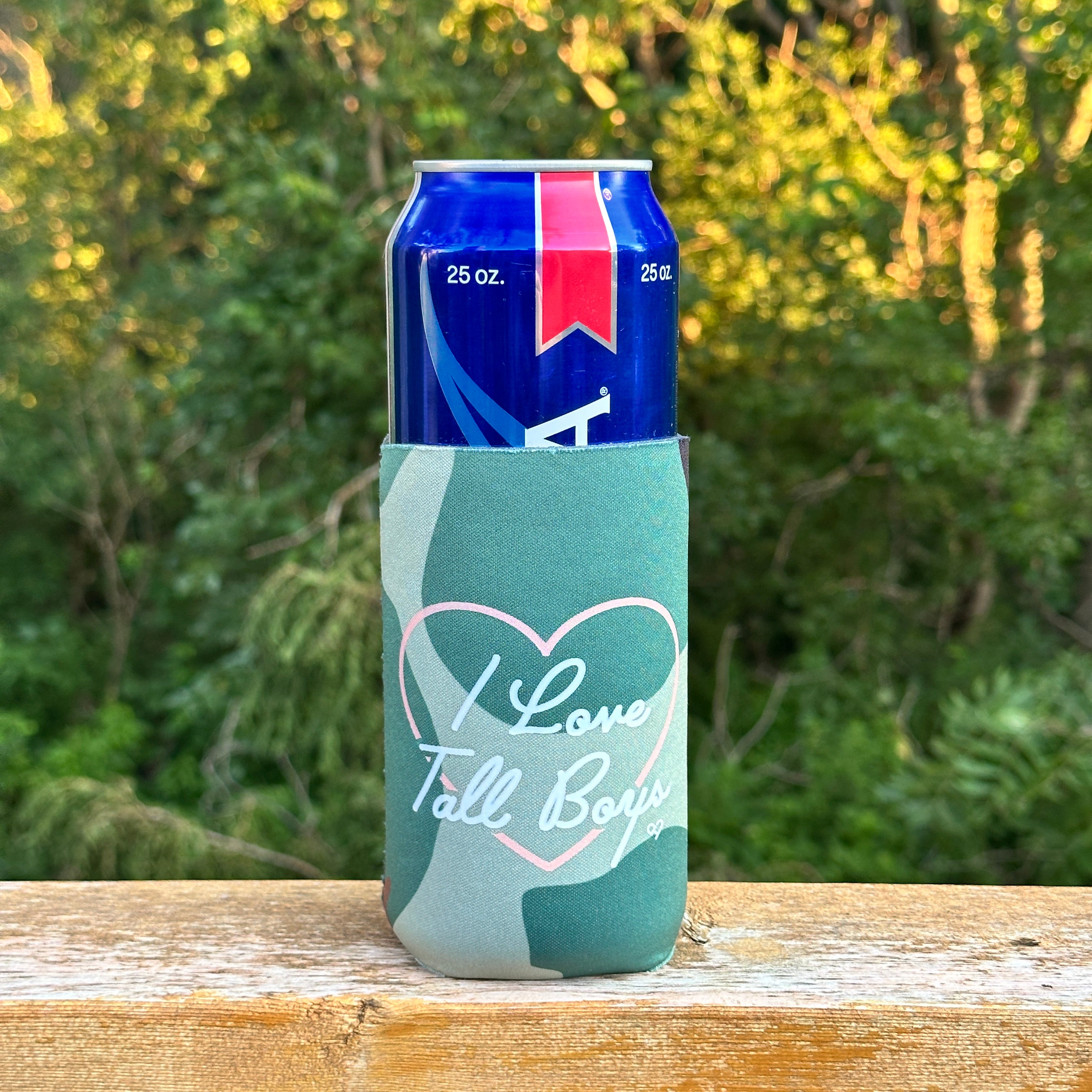 I Love Tall Boys Camo Koozie, green camo slim sleeve with pink heart and white script on a tall can on a railing