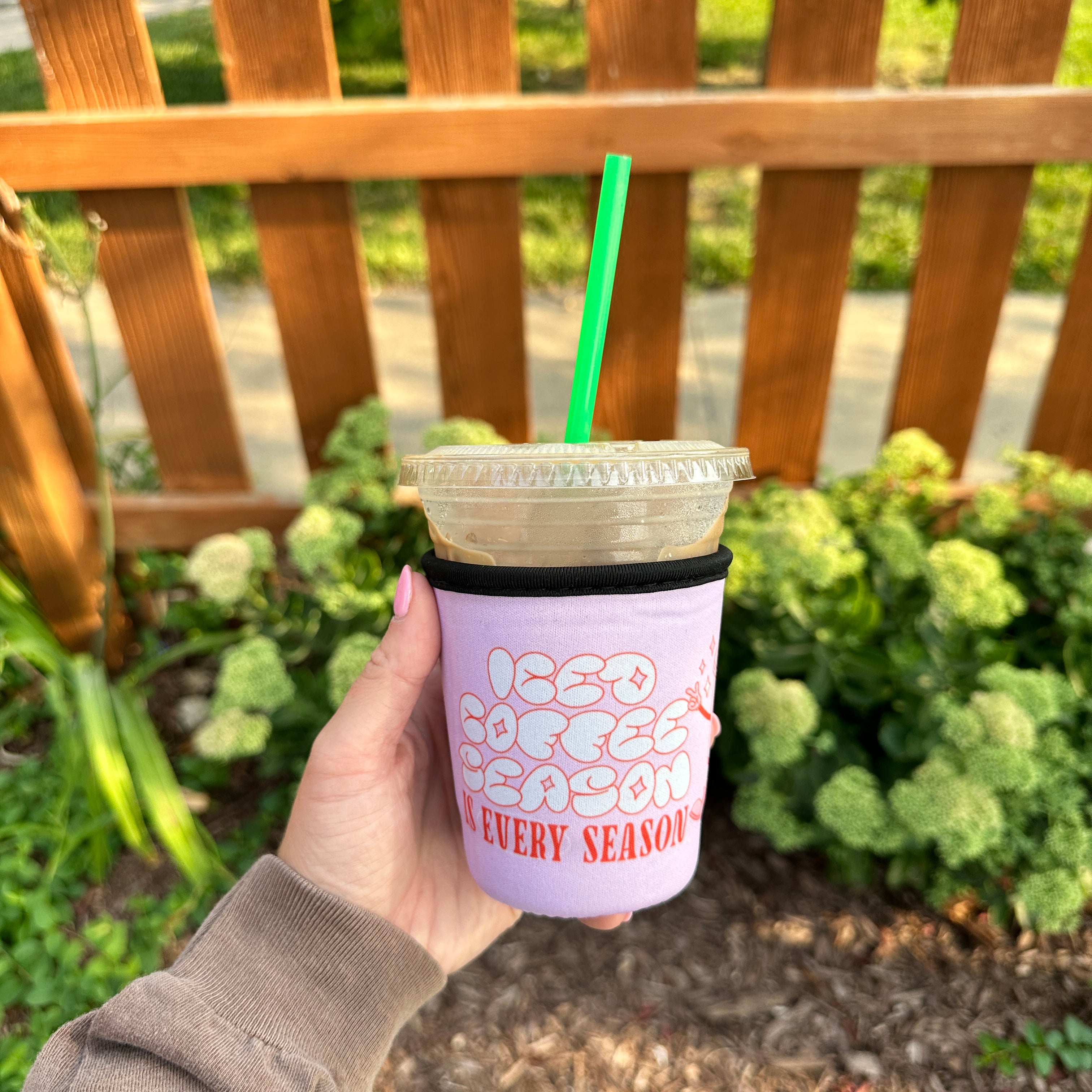 Iced Coffee Season Is Every Season Sleeve, lavender sleeve on an iced coffee with green straw held by a wooden fence