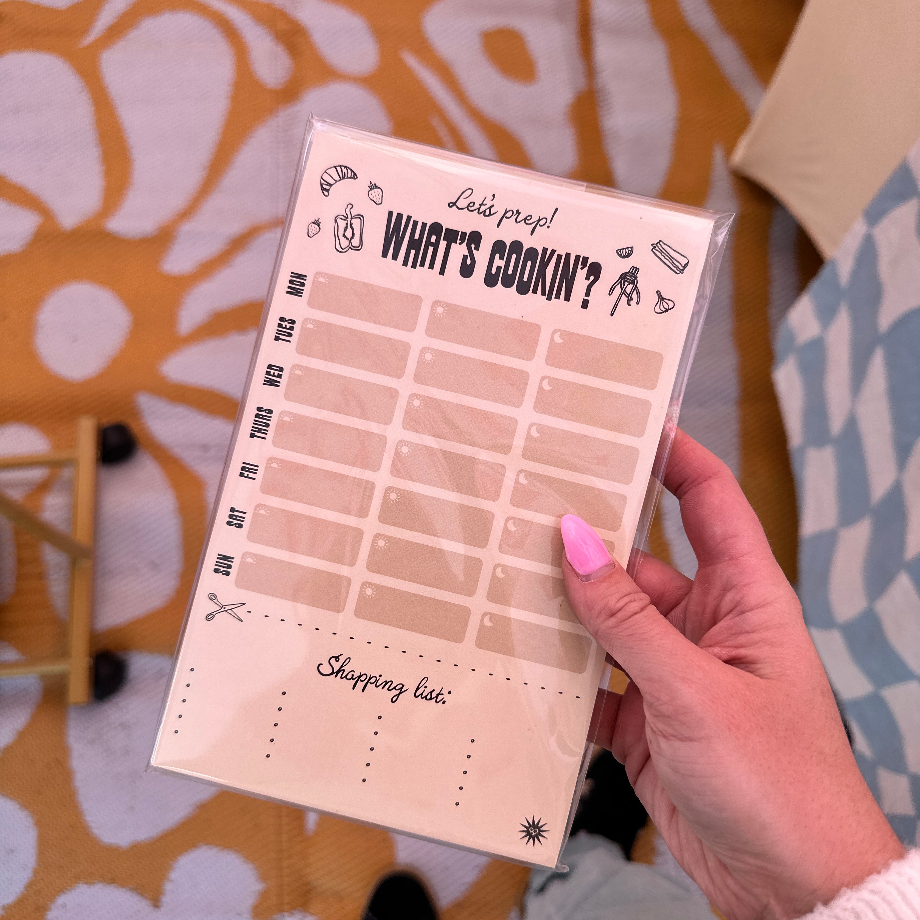 What's Cookin' Meal Prep Pad, cream weekly meal planner with shopping list, held in hand over a yellow floral rug
