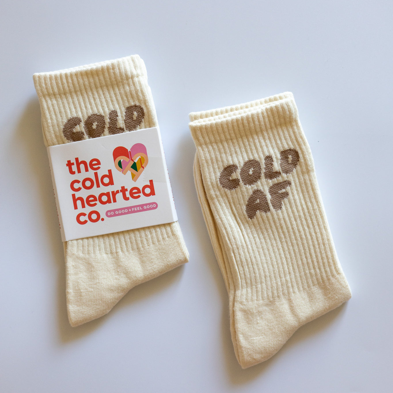 Cold AF Socks, two cream crew socks with brown Cold AF lettering, one in a Cold Hearted Co. band, on a white surface