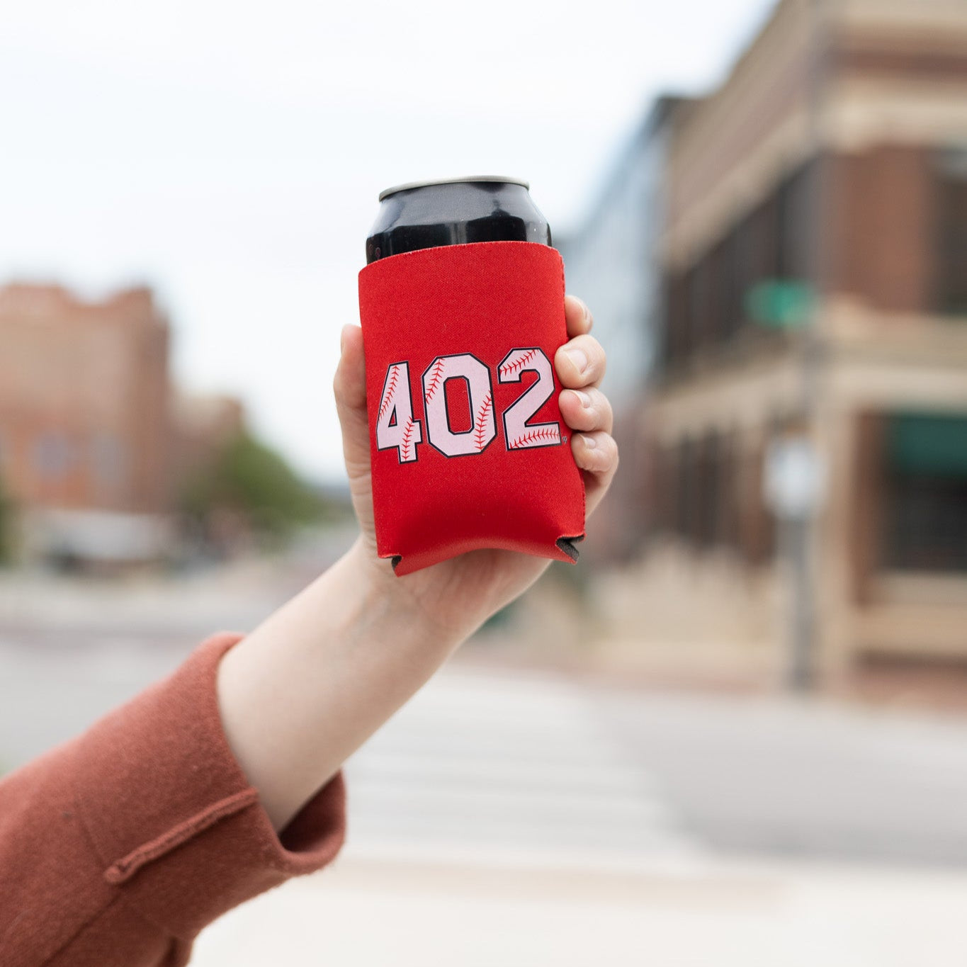 402 Baseball Koozie, hand holding a red koozie with white baseball stitch 402 numbers on a city street
