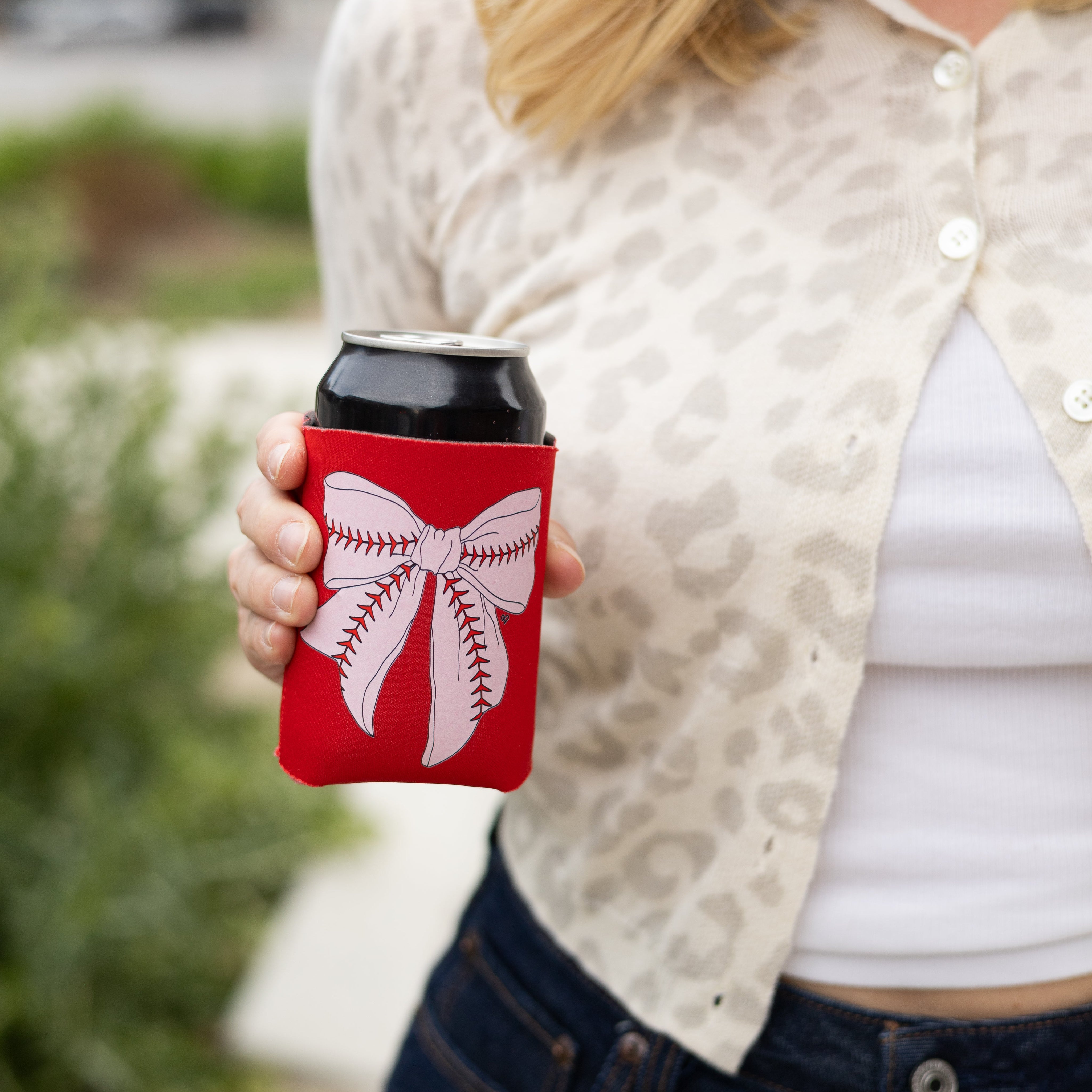 Baseball Bow Koozie held by a model in a leopard cardigan, red koozie with white baseball bow graphic on a can