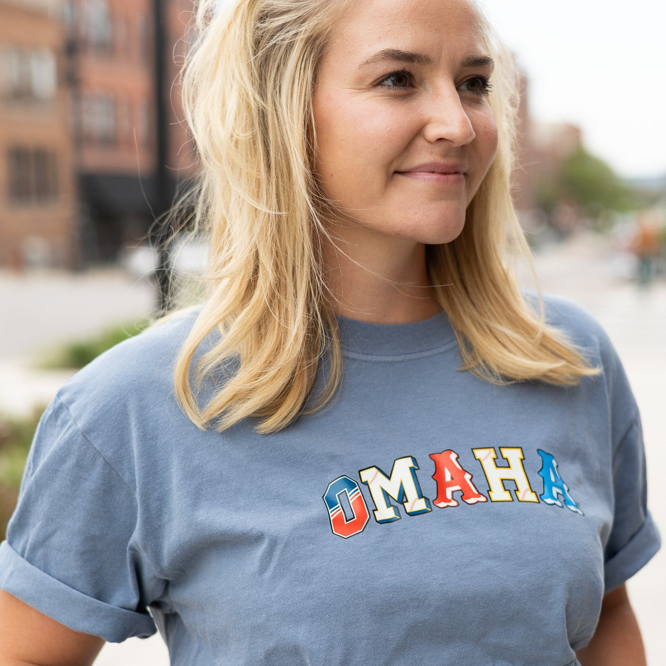 Omaha Baseball Tee worn by a smiling model on a downtown street, blue tee with multicolor arched OMAHA letters