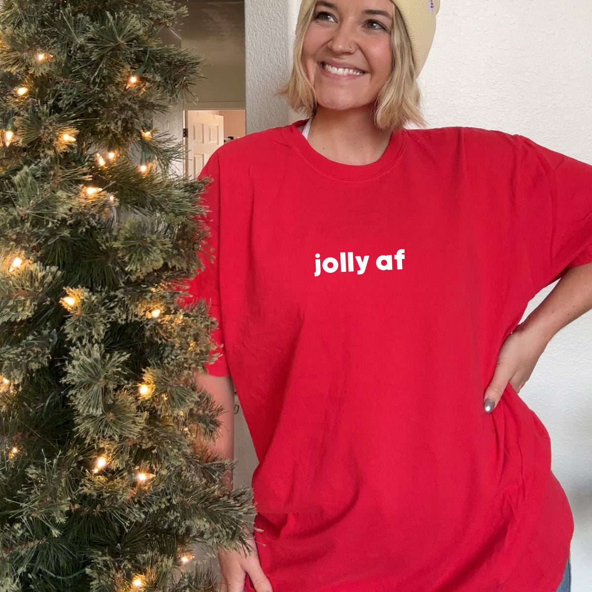 Jolly AF Tee in red worn oversized by a model in a cream Holidays beanie beside a lit Christmas tree