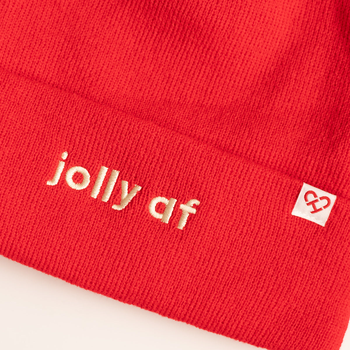Jolly AF Beanie, close up of red knit cuff with cream embroidered jolly af text and a white heart logo tag