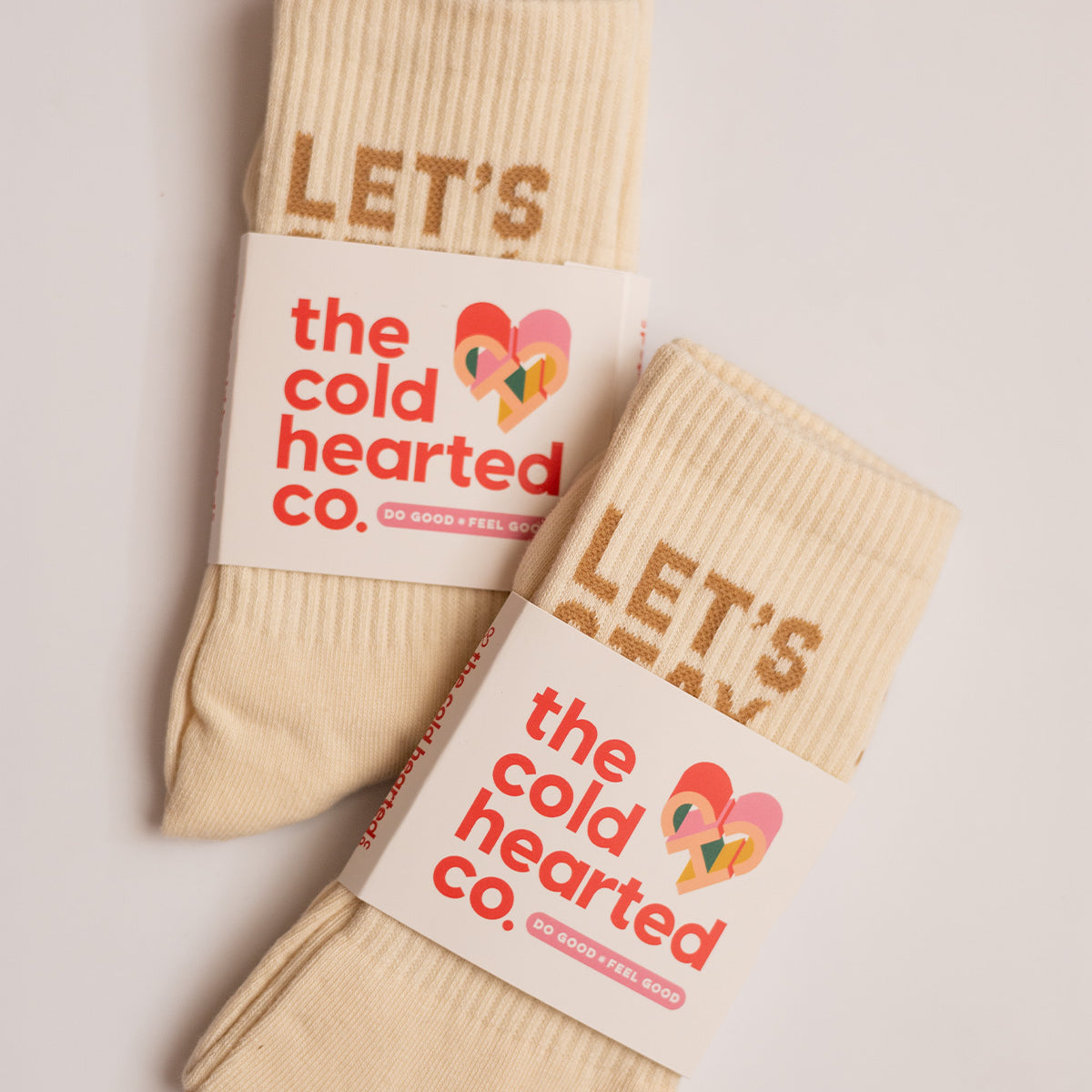 Let's Stay Home Socks, two folded cream crew socks with tan lettering in The Cold Hearted Co. paper bands