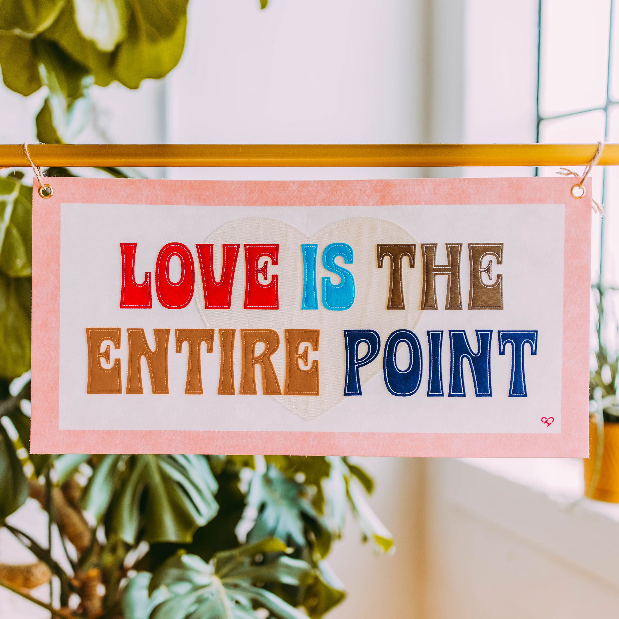 Love is The Entire Point Pennant, pink bordered felt banner in red, blue and brown lettering hung by a window