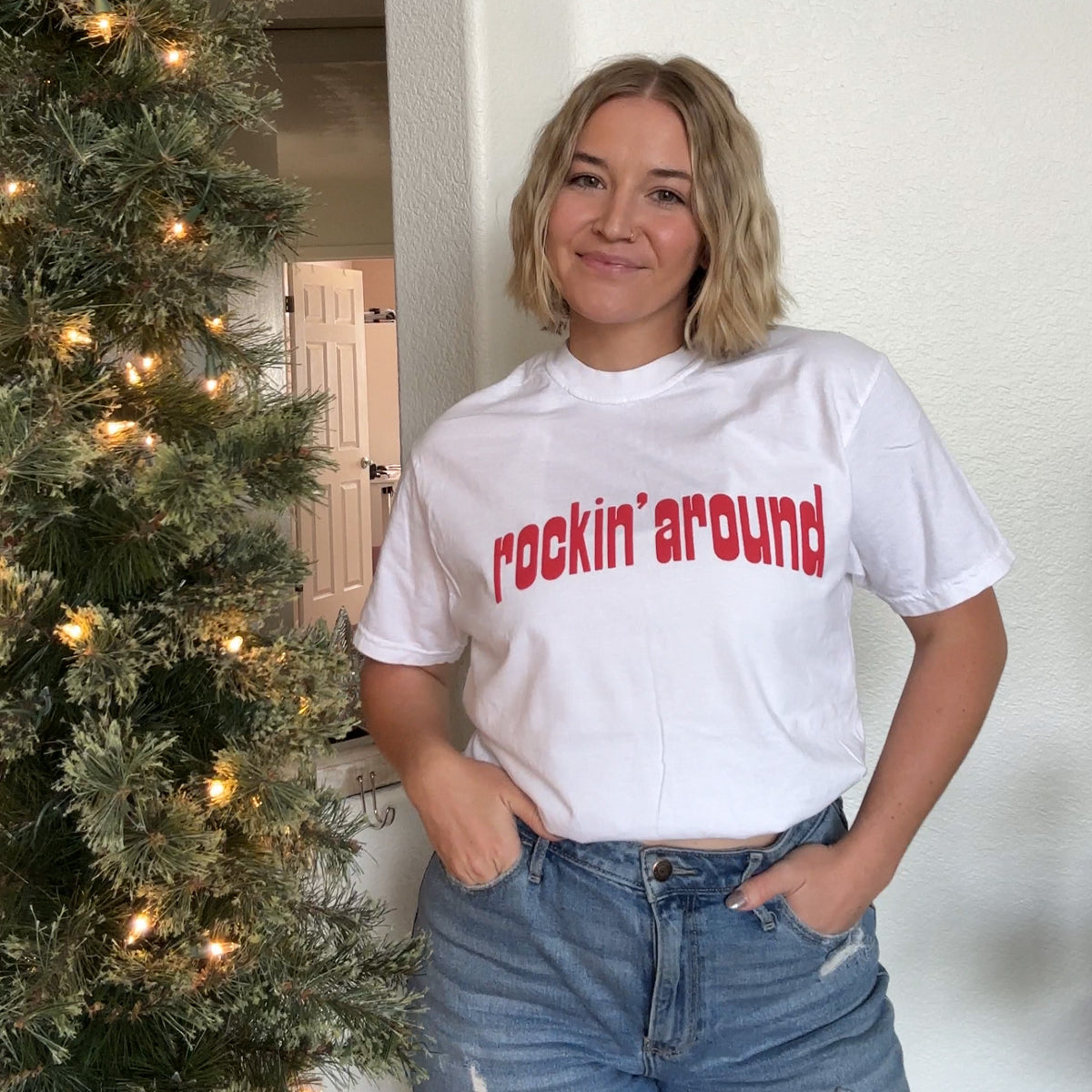 Rockin' Around Tee in white with red text worn by a smiling model in jeans beside a lit Christmas tree