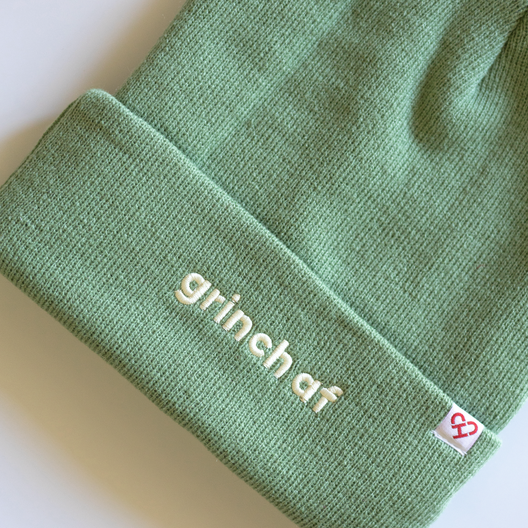 Grinch AF Beanie, close up of sage green knit cuff with raised cream grinch af embroidery and red heart logo tag