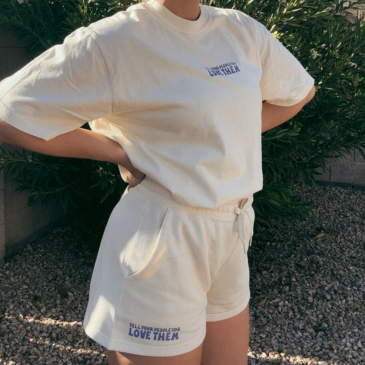 Tell Your People You Love Them Sweat Shorts in cream, worn by a model with a matching tee against green shrubs