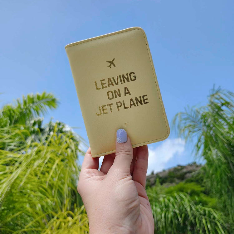 Leaving On A Jet Plan Passport Wallet in pale yellow with gold foil text and plane icon held up by palm trees